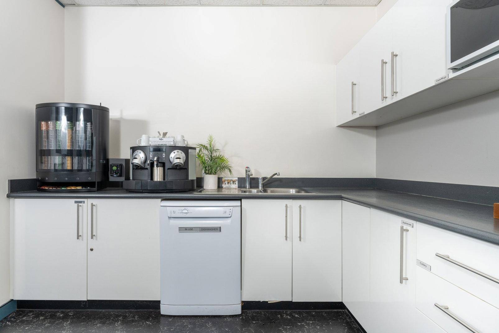 Funktionale Büroküche mit weißen Schränken, Spülmaschine, Kaffeemaschine und Verkaufsautomat.