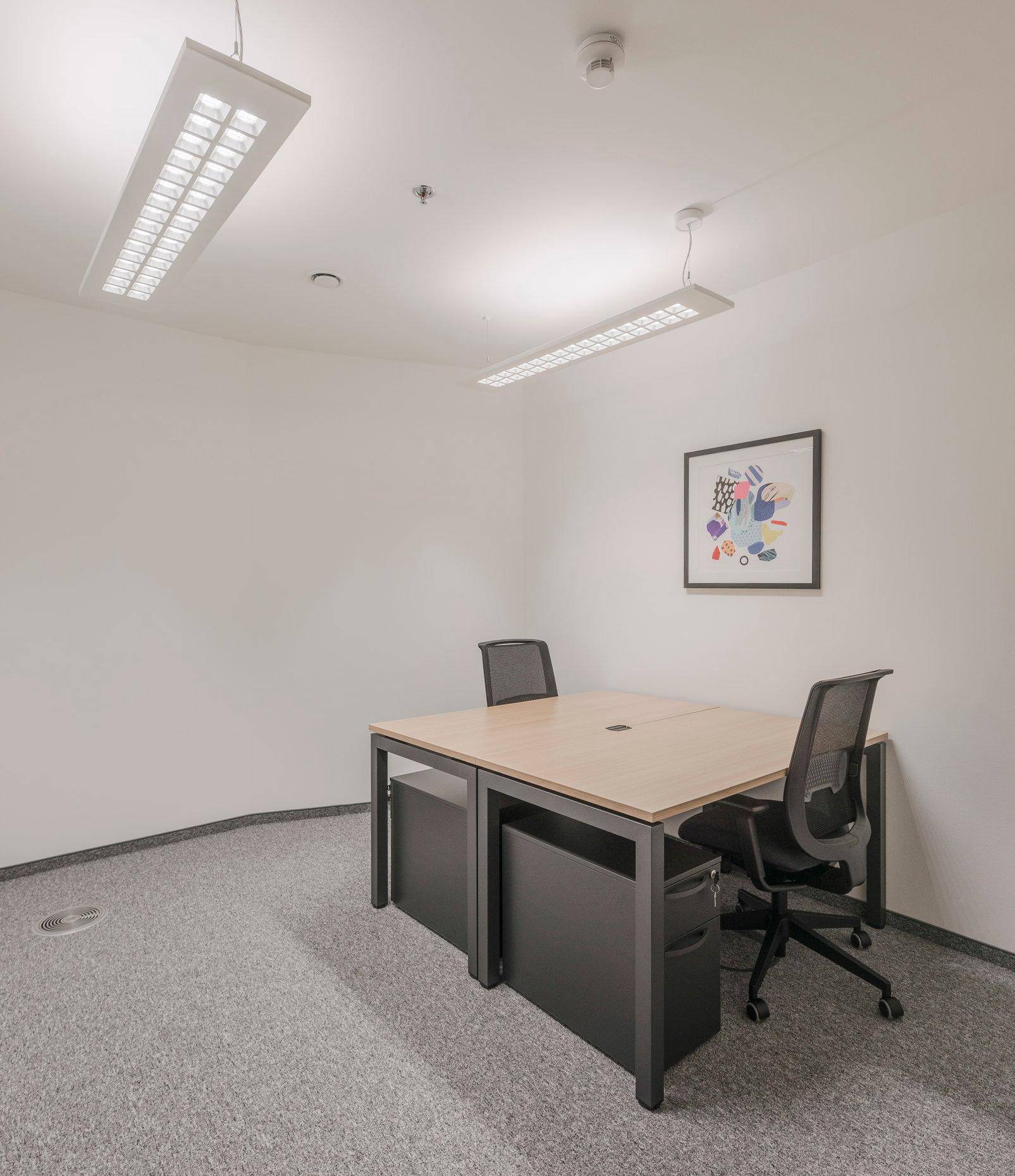 Modern eingerichtetes Büro mit einem Doppelarbeitsplatz, Teppichboden und heller Deckenbeleuchtung.