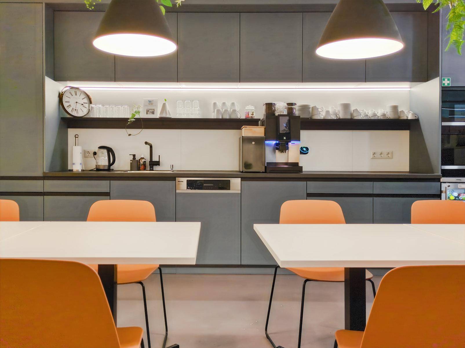 Moderne Teeküche mit grauen Schränken, Kaffeemaschine, Spülmaschine und orangefarbenen Stühlen für eine lebendige Atmosphäre.