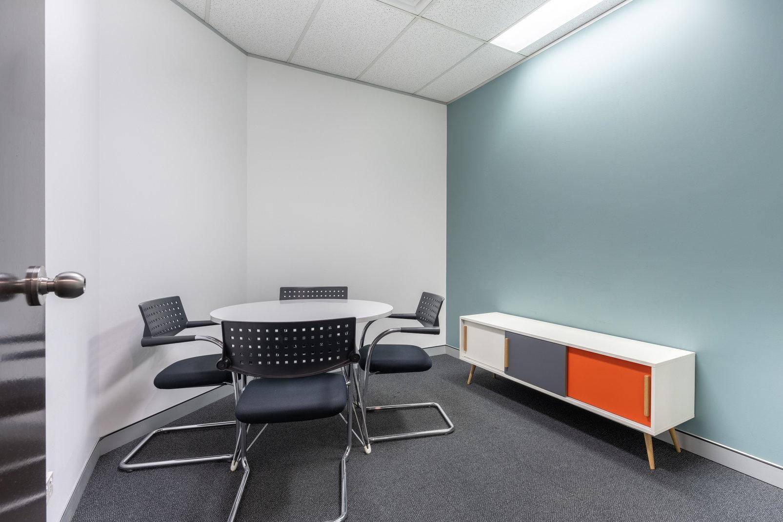 Kleiner Besprechungsraum mit rundem Tisch, vier Stühlen und einem farbigen Sideboard für effiziente Meetings.
