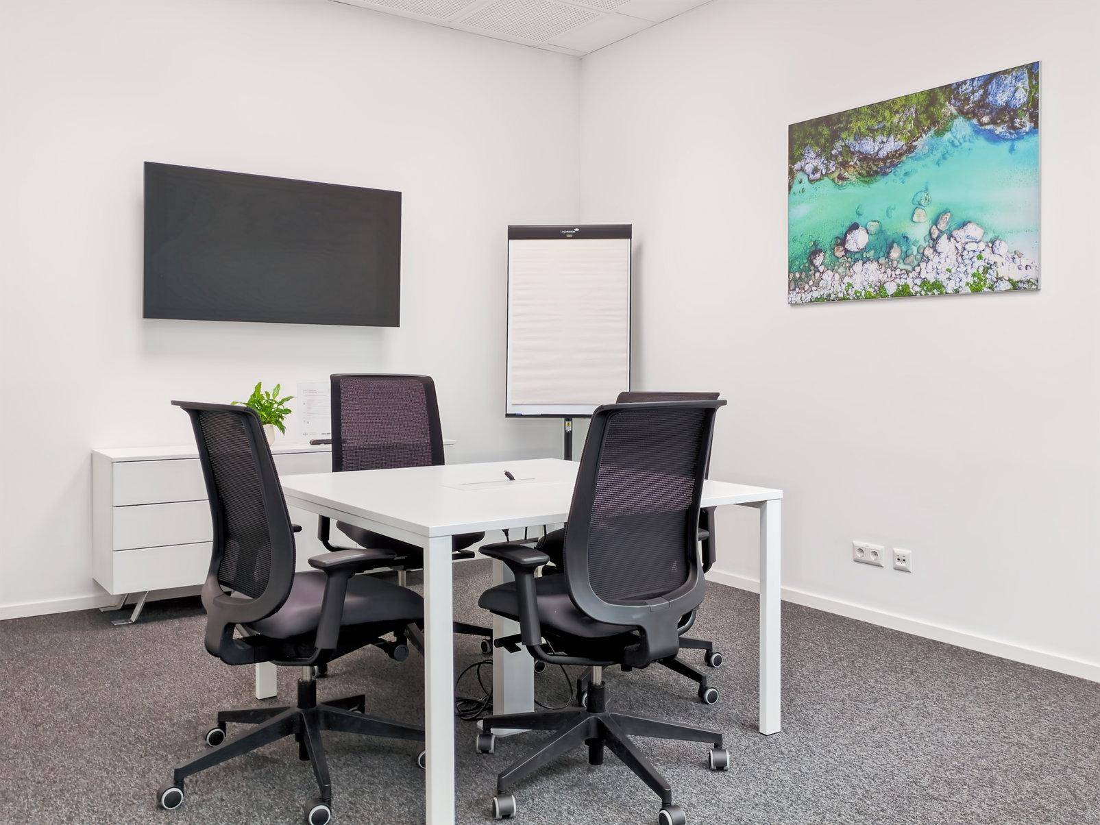 Kleiner Besprechungsraum mit weißem Tisch, vier Stühlen, Wandbildschirm und Flipchart für Präsentationen.