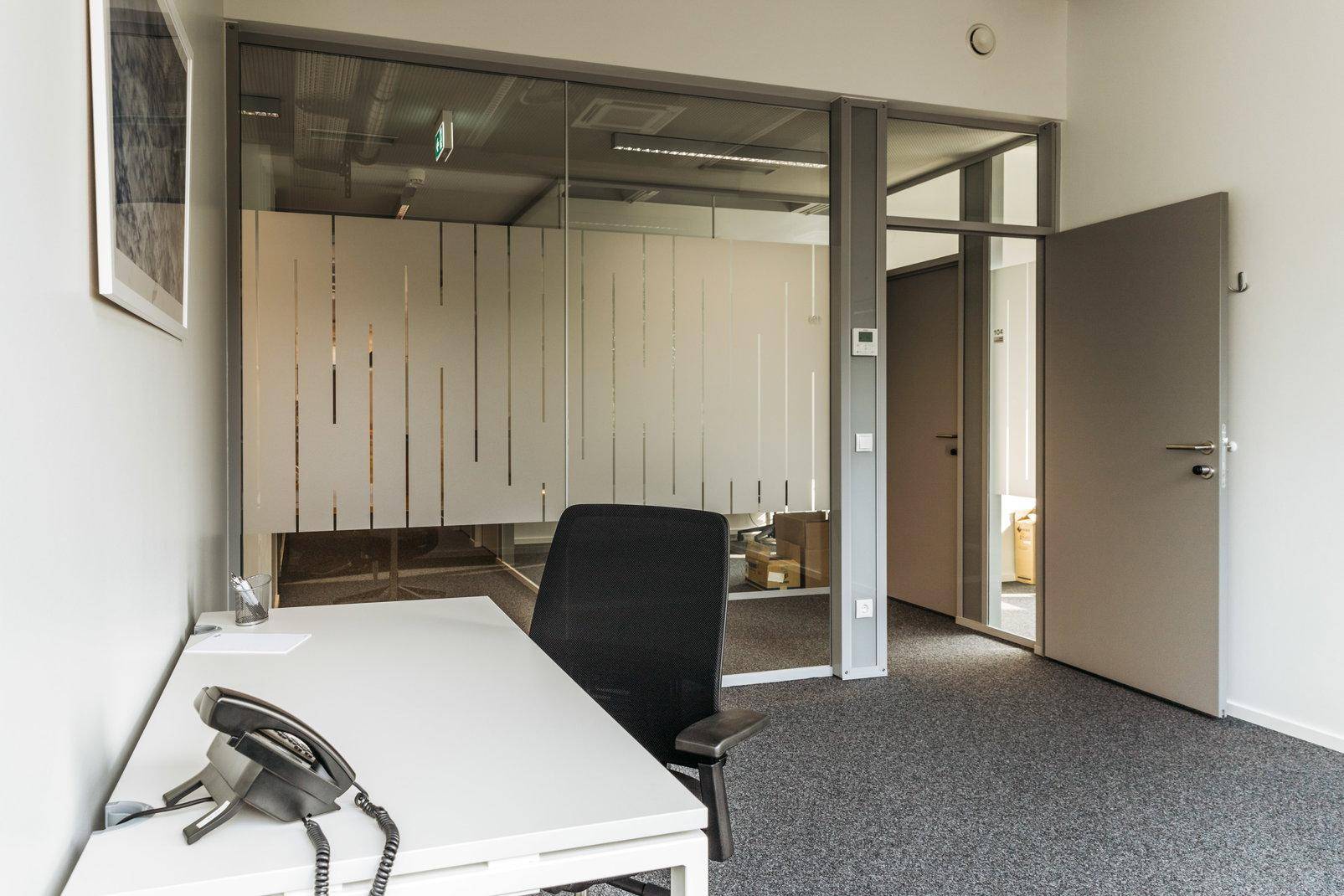 Modernes Einzelbüro mit Teppichboden, Schreibtisch und Glaswand zu einem weiteren Raum.
