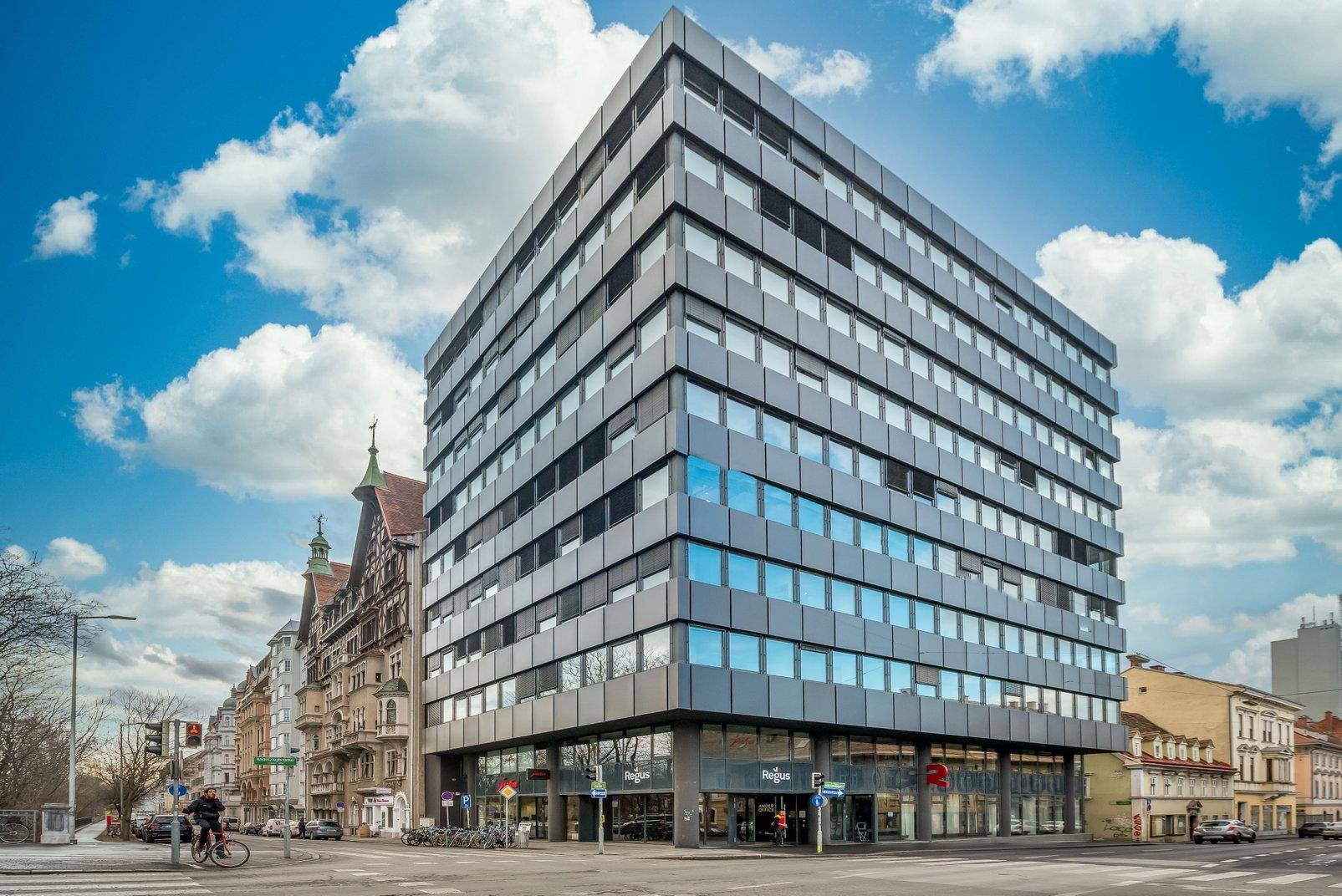 Modernes Bürogebäude an einer Straßenecke mit markanter Glas- und Metallfassade unter blauem Himmel.