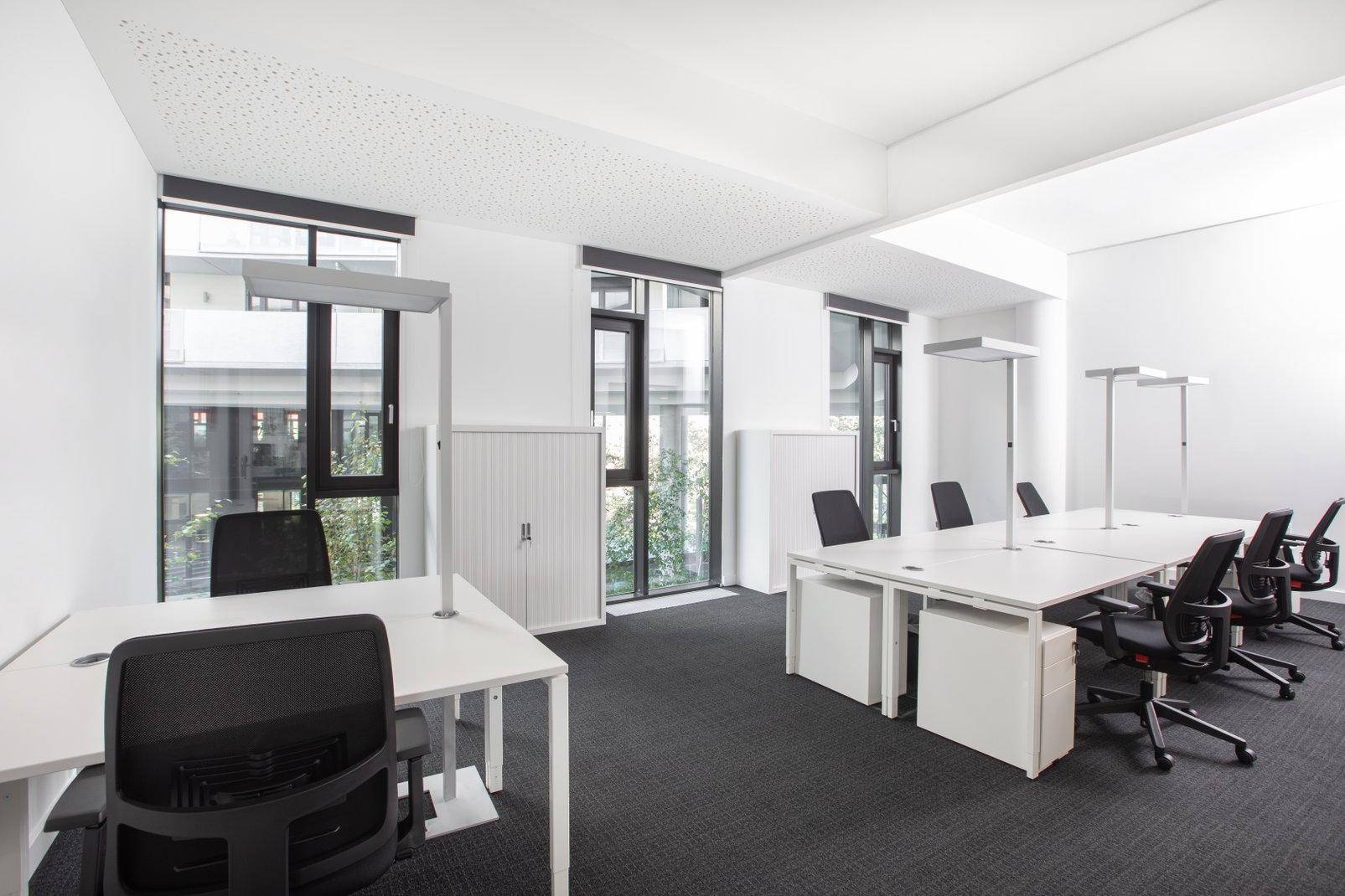 Helles Büro mit mehreren Arbeitsplätzen, Teppichboden und großen Fenstern mit Blick ins Grüne.
