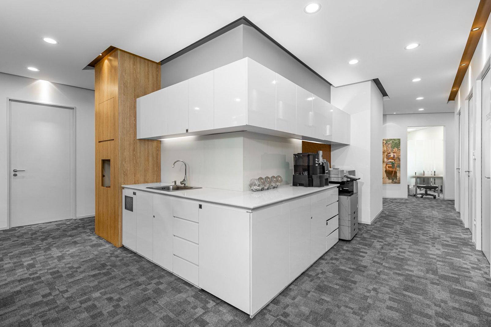 Moderne Teeküche mit weißen Hochglanzschränken, Spüle, Kaffeemaschine und Teppichboden in einem Büroflur.