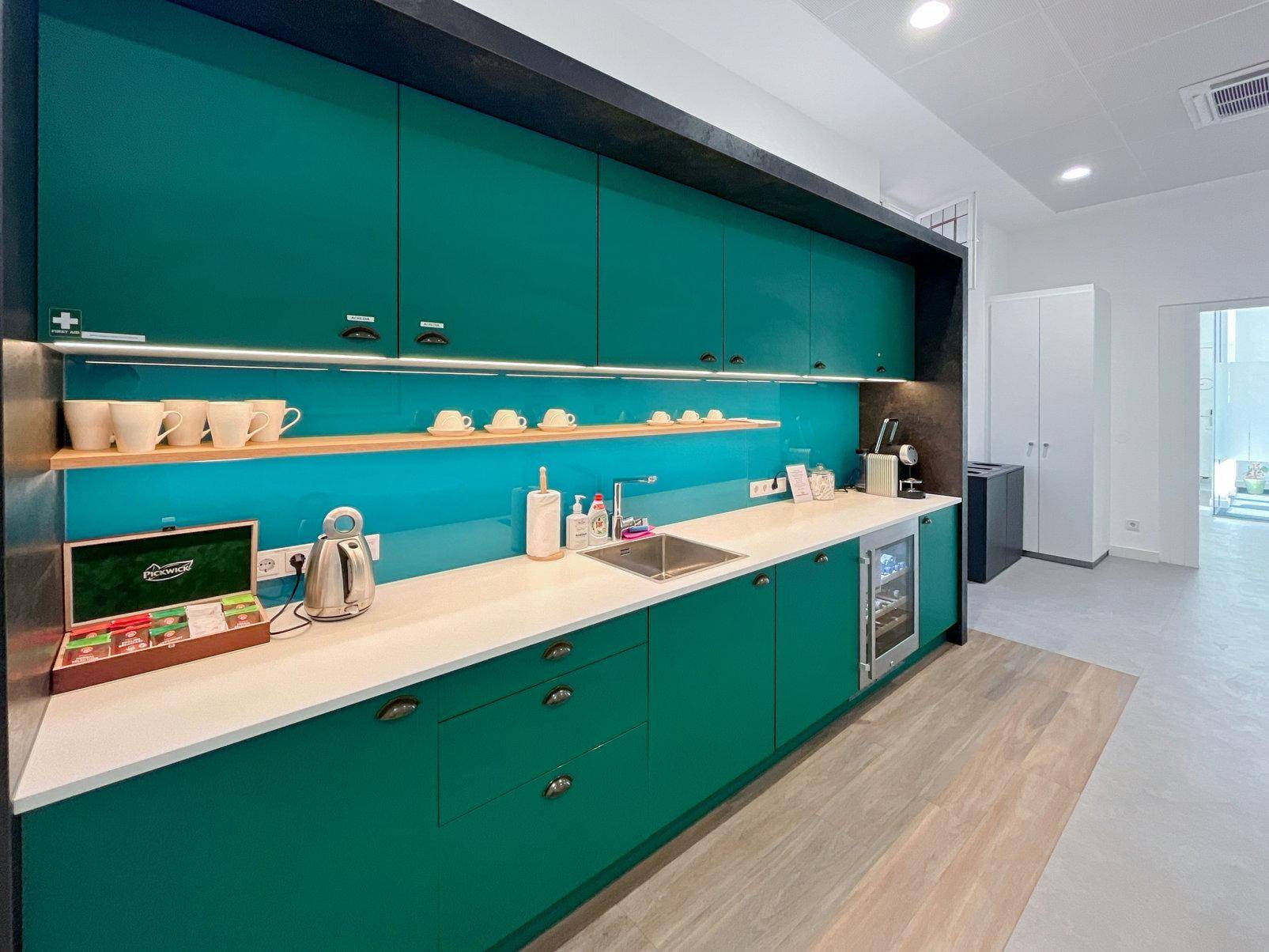 Moderne Teeküche mit teal-farbenen Schränken, weißer Arbeitsplatte und integriertem Kühlschrank.