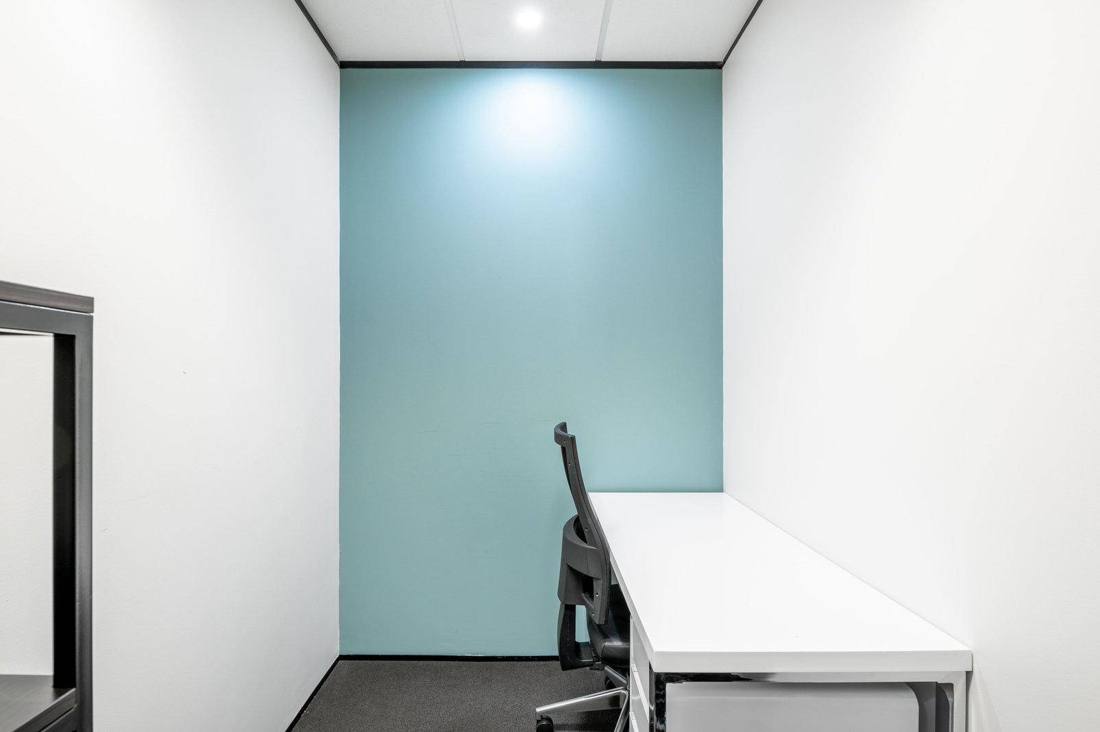 Schmaler Büroraum mit einem langen weißen Schreibtisch, Bürostuhl und einer hellblauen Akzentwand.