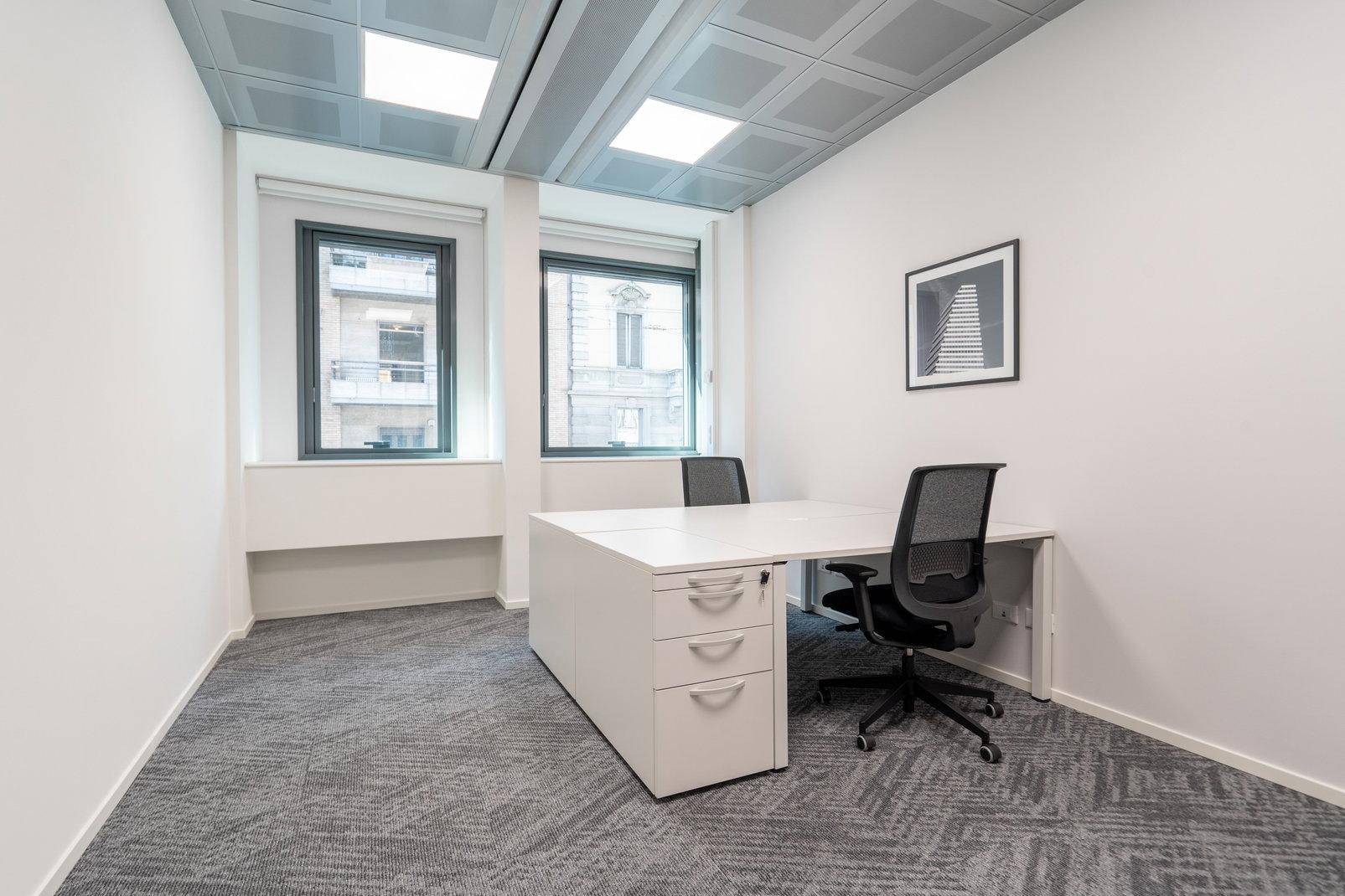 Kleines, modernes Büro mit weißem Schreibtisch, zwei Bürostühlen und Teppichboden in Grau.