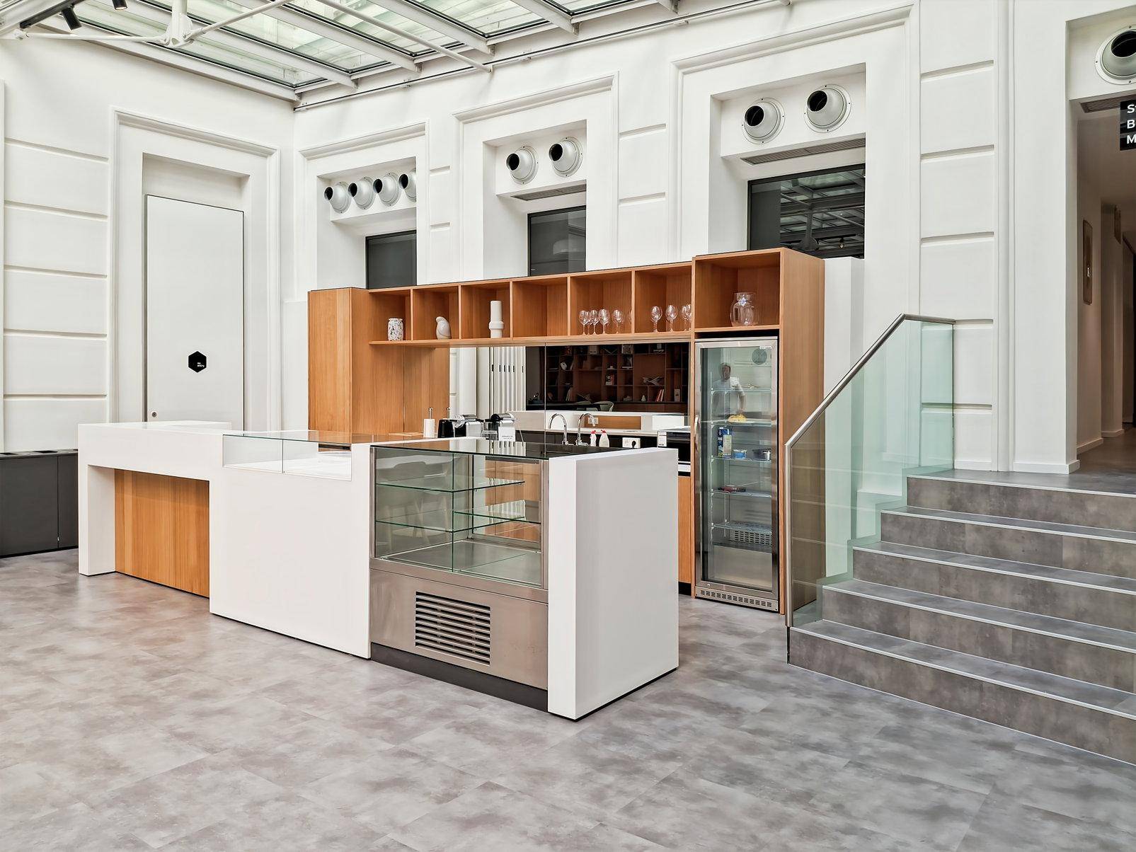Heller Café- und Barbereich mit Glasvitrine, Holzregalen, Kühlschrank und Treppe unter einem Glasdach.