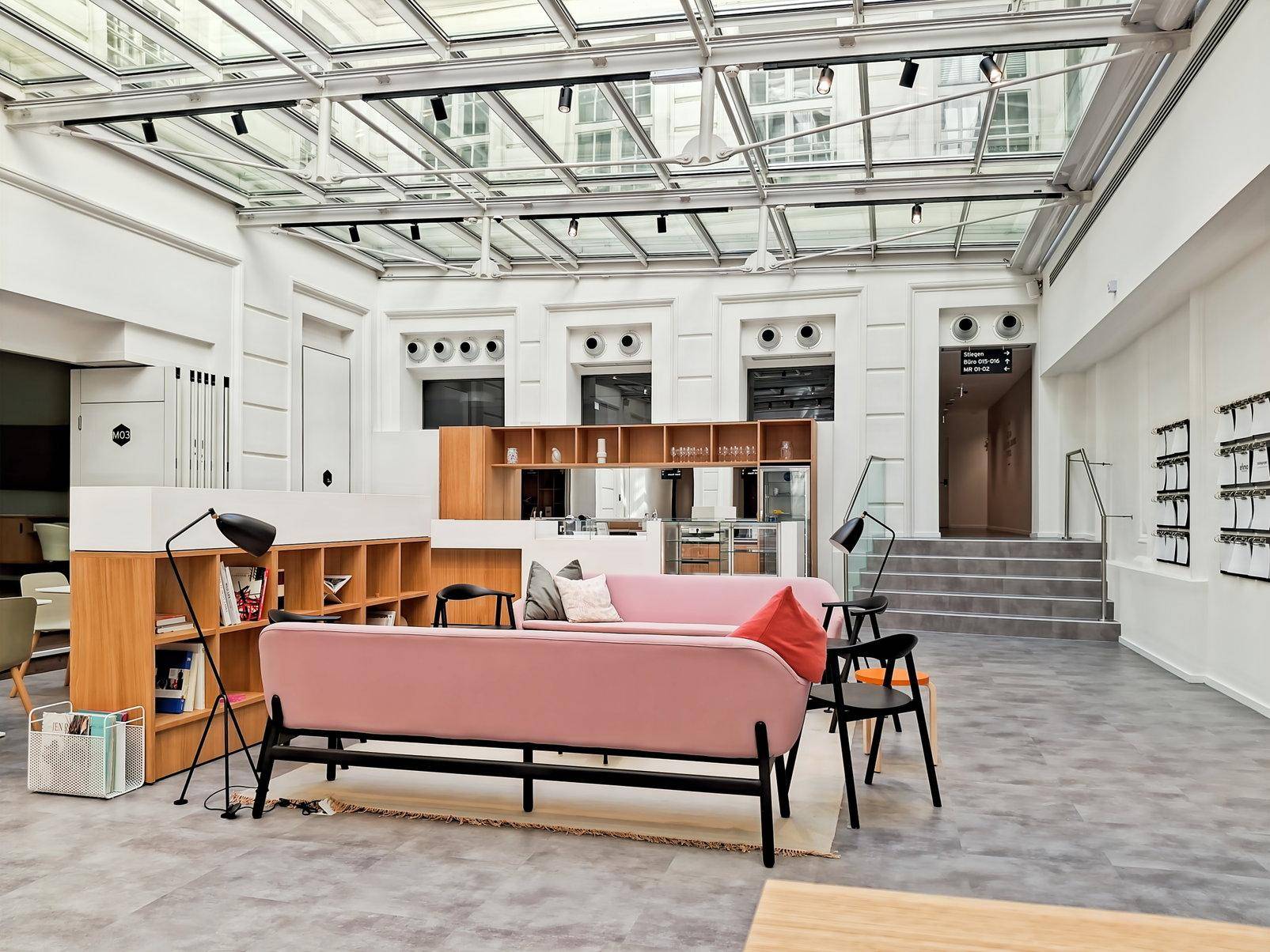 Heller Lounge-Bereich mit Glasdach, moderner Einrichtung, rosa Sofa und Holzregalen.
