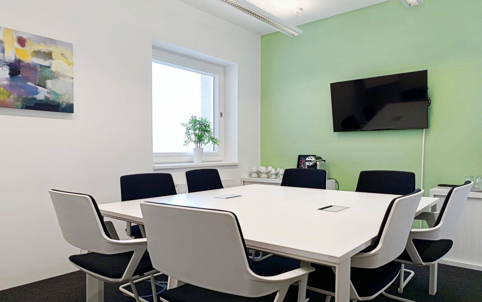 Heller Besprechungsraum mit weißem Tisch, modernen Stühlen, einem Fenster und einer hellgrünen Akzentwand.