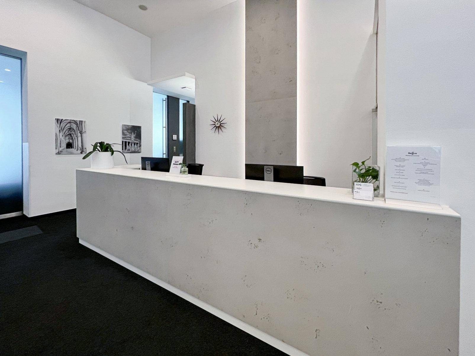 Empfangsbereich mit einer langen, hellen Theke, zwei Monitoren und minimalistischer Dekoration auf dunklem Teppichboden.