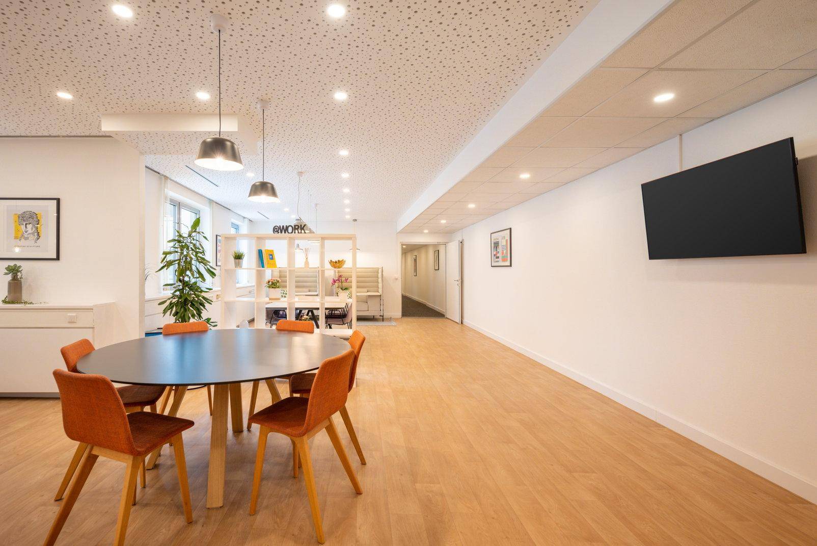 Geräumiger Loungebereich mit Holzboden, runden Tischen und einem Flachbildfernseher an der Wand.