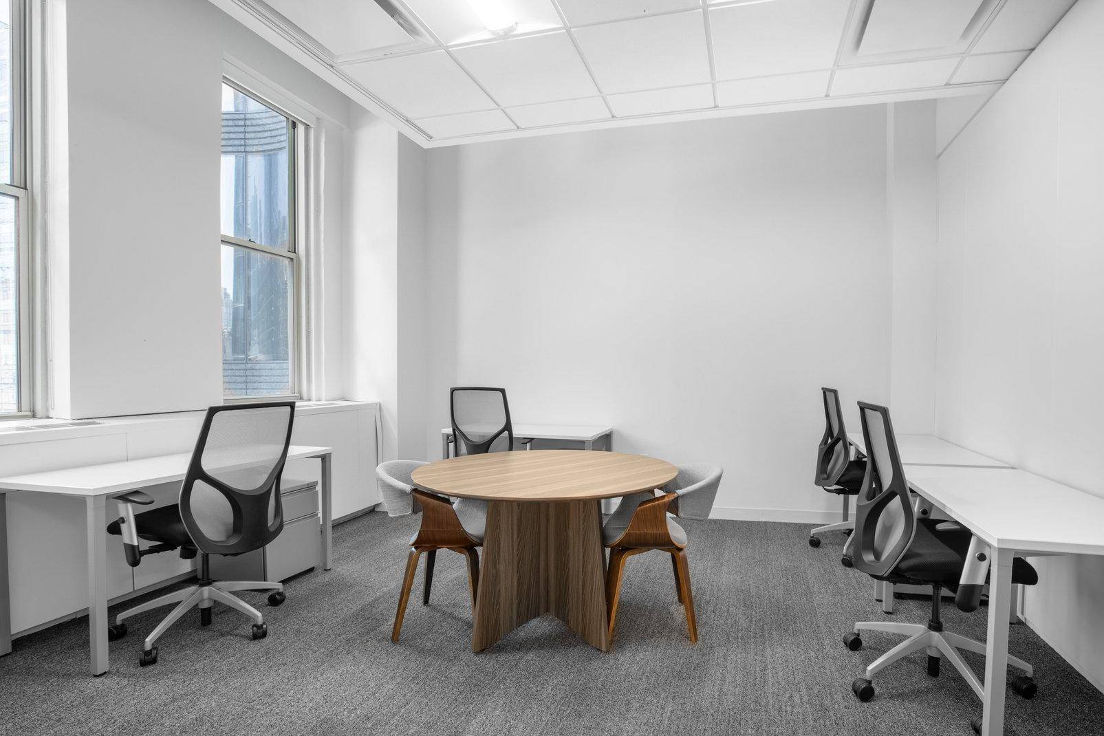 Kleines Büro mit mehreren Arbeitsplätzen, einem runden Besprechungstisch und einem großen Fenster für Tageslicht.