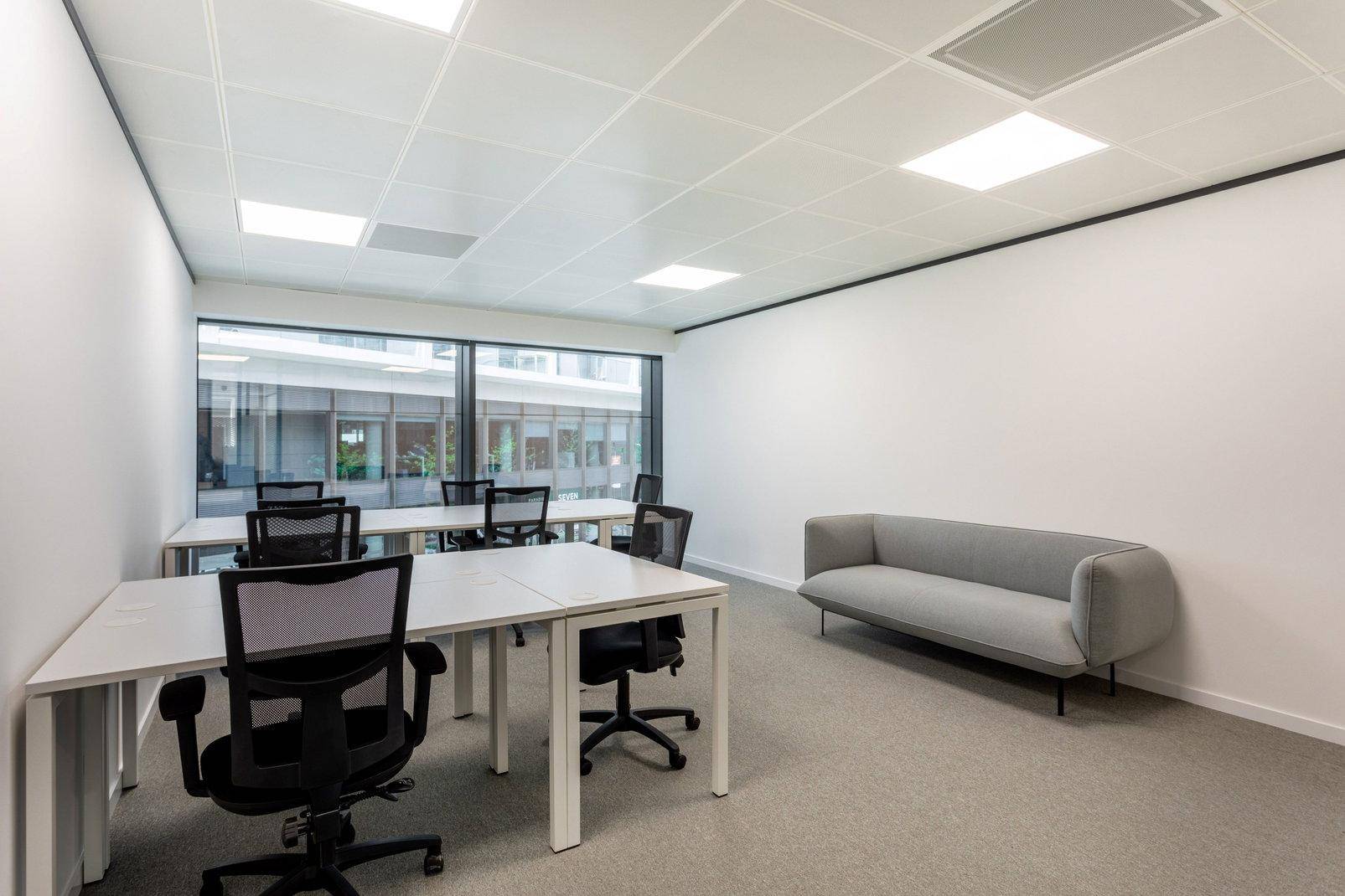 Modernes Büro mit mehreren Schreibtischen, Bürostühlen und einem Sofa, mit Blick auf einen Innenhof.