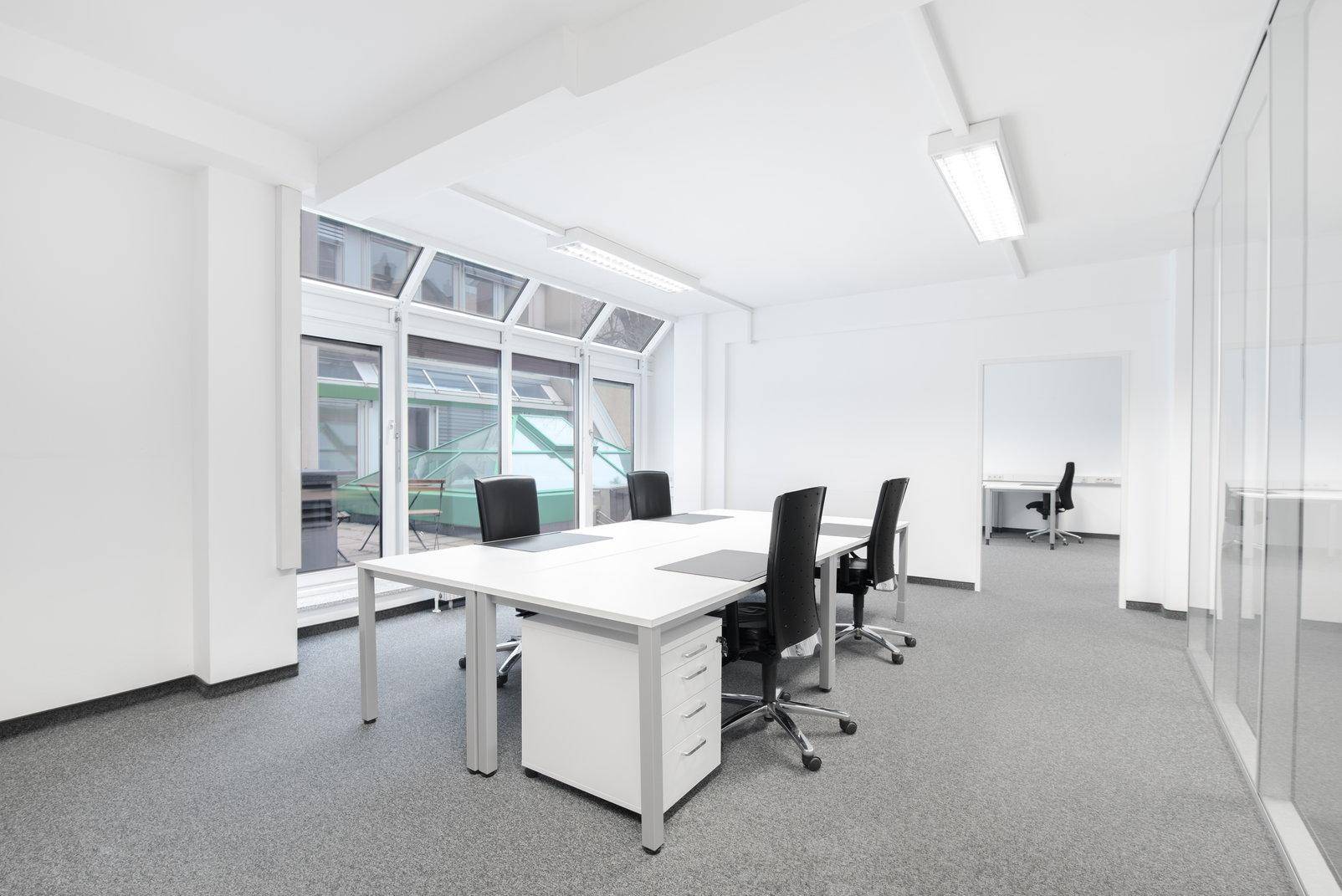 Helles Büro mit mehreren Schreibtischen, Teppichboden und großen Fenstern mit Blick auf einen Innenhof.
