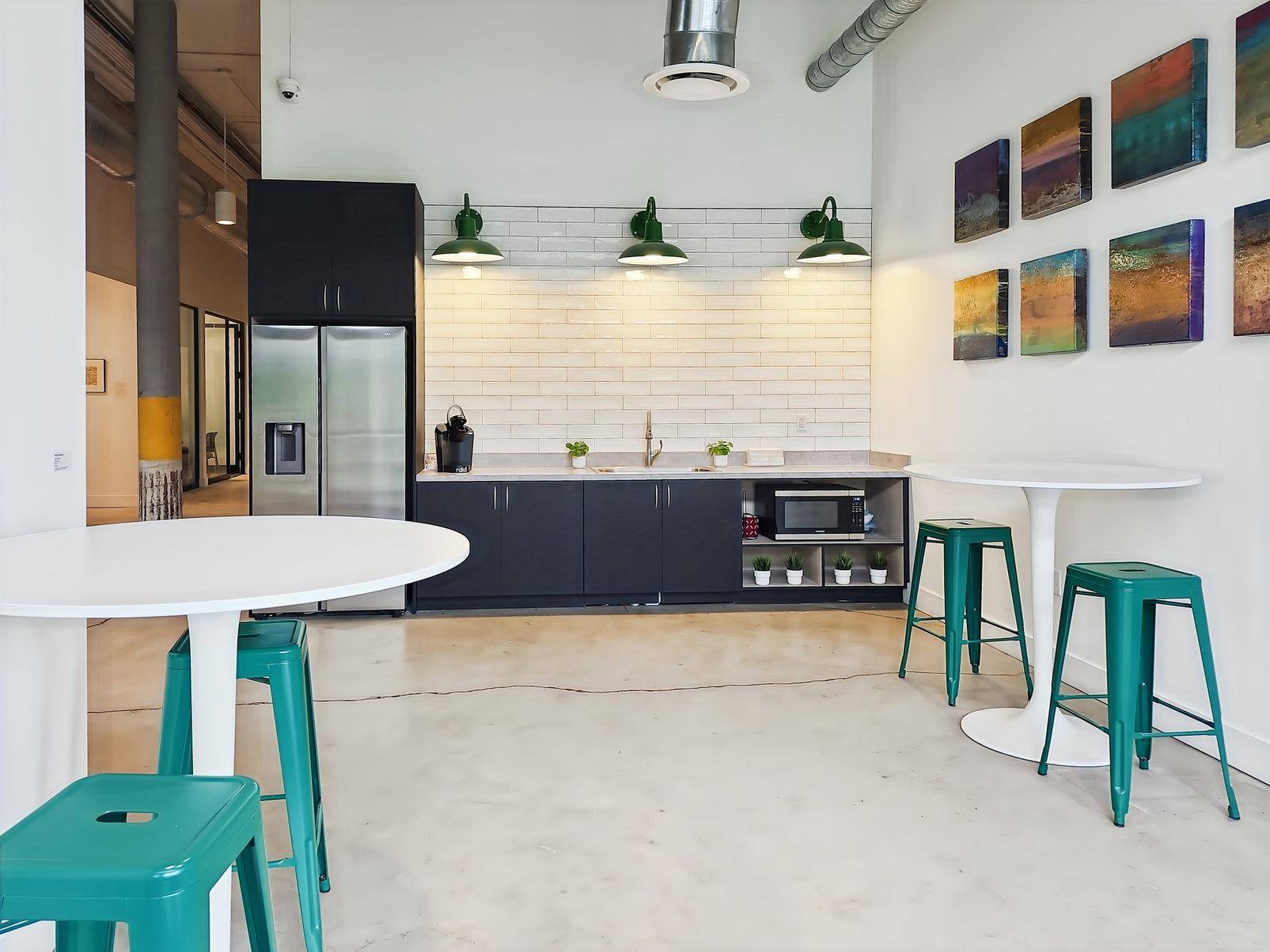 Moderne Teeküche mit dunklen Schränken, weißer Fliesenrückwand und Stehtischen mit Hockern.