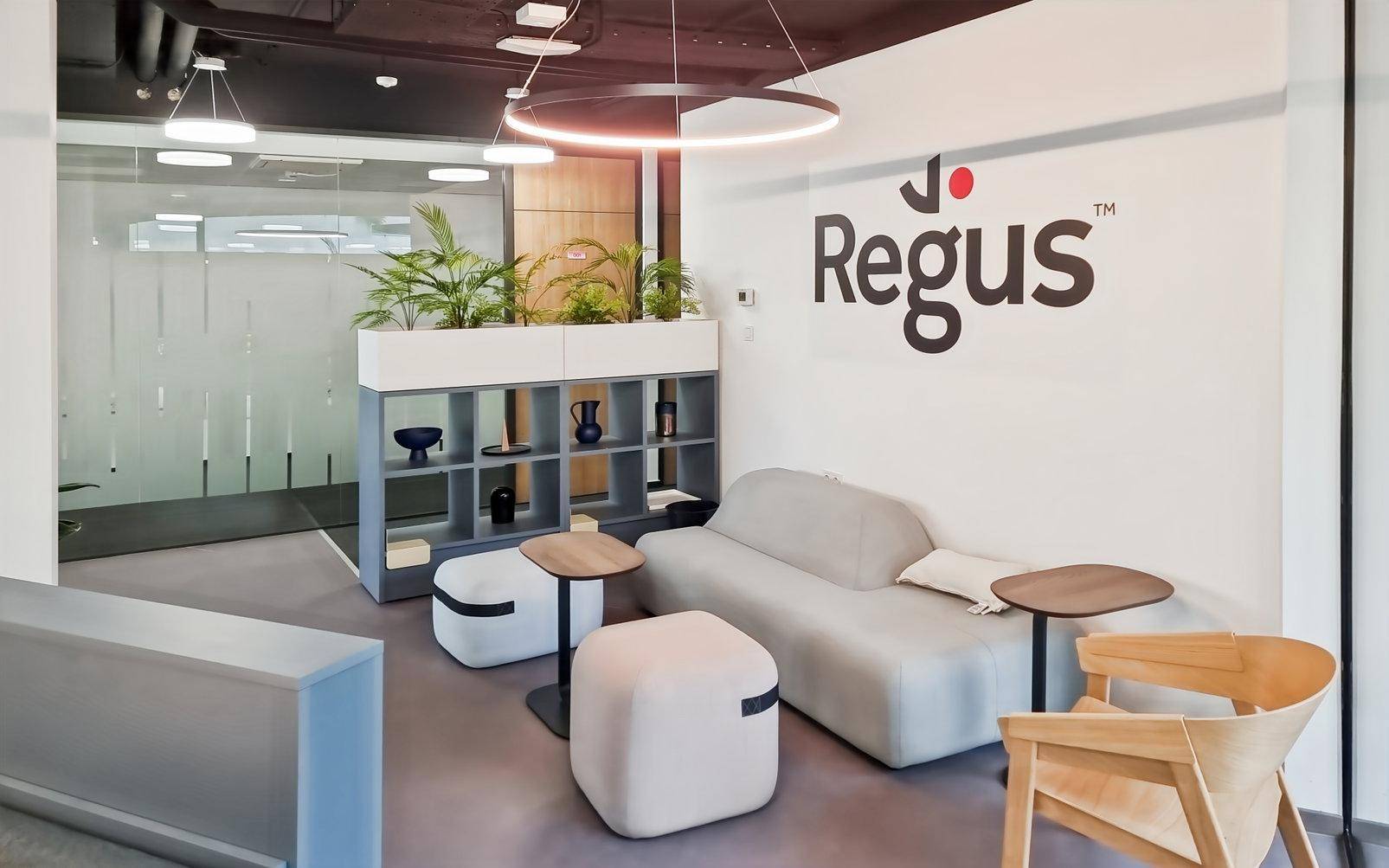 Gemütlicher Loungebereich mit modernen Sitzgelegenheiten, Pflanzen und dem Regus-Logo an der Wand, ideal zum Entspannen.