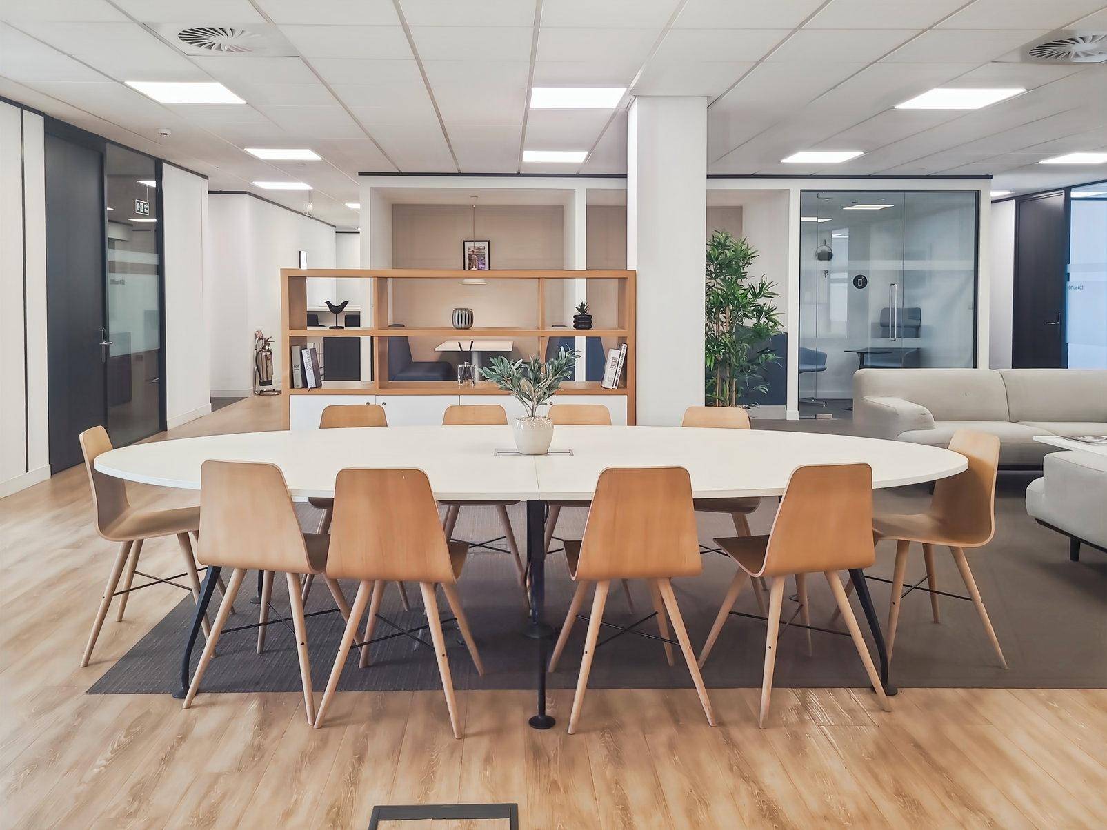 Moderner Bürobereich mit großem ovalen Tisch, Holzstühlen und offener Gestaltung mit Regalen und Sitzgelegenheiten.