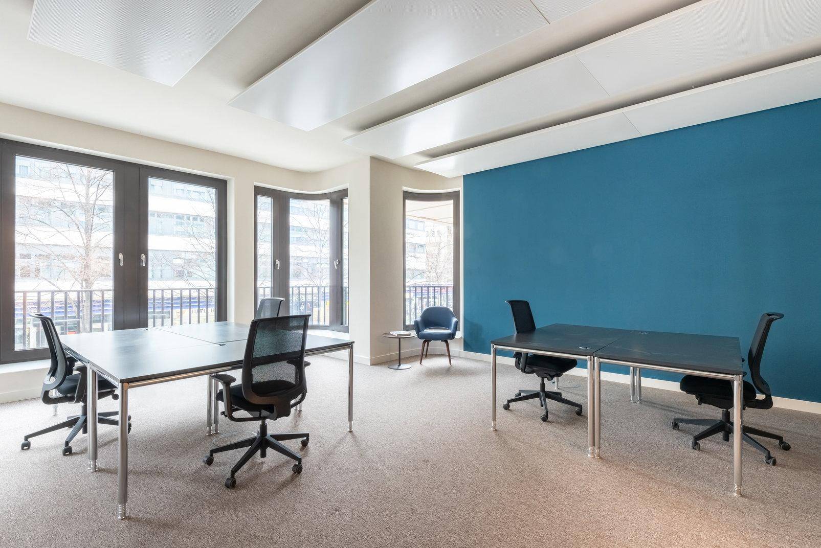 Helles Büro mit blauer Akzentwand, mehreren Arbeitsplätzen und großen Fenstern.