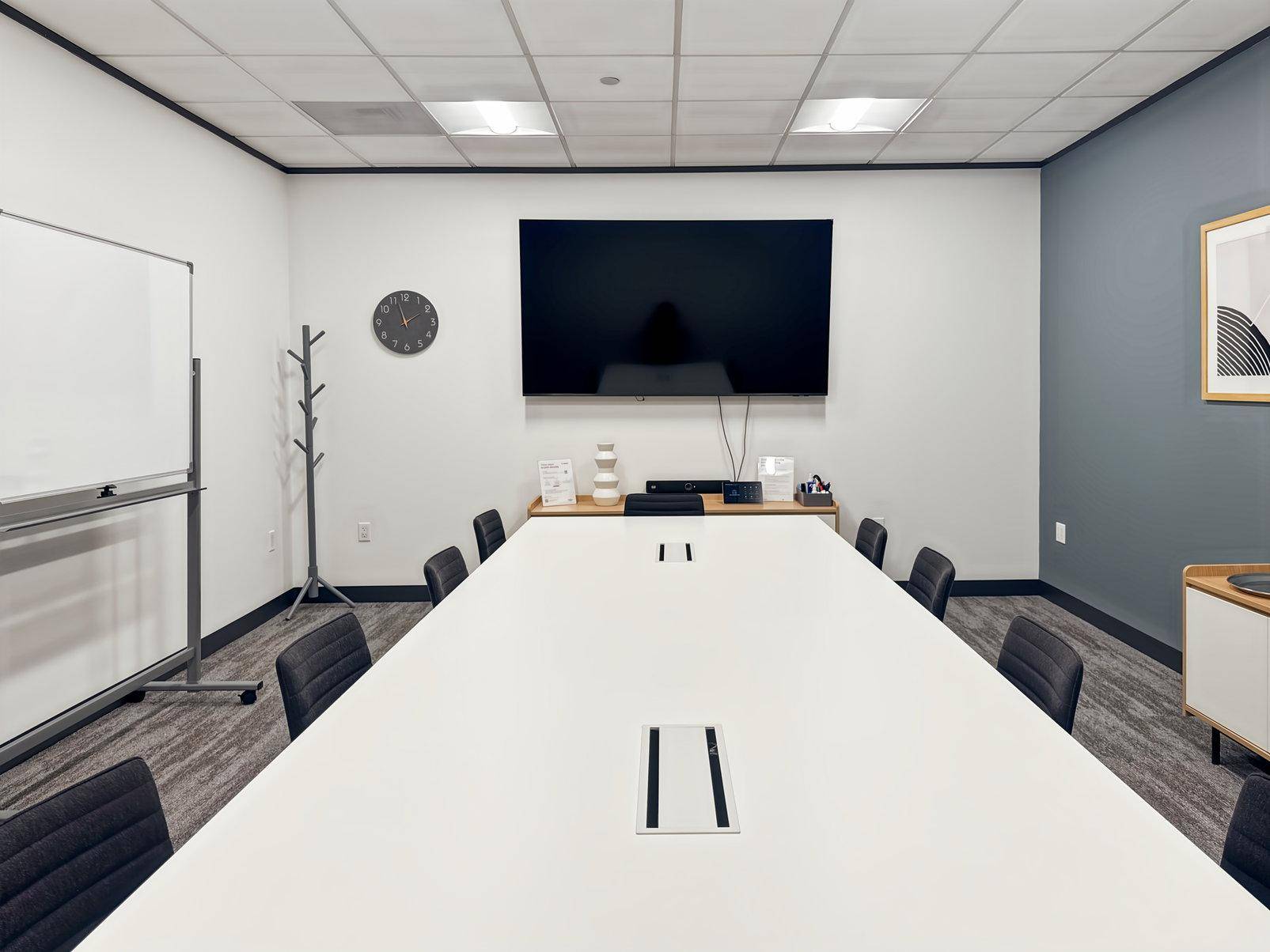 Konferenzraum mit großem Tisch, Wandfernseher, Whiteboard und Teppichboden.