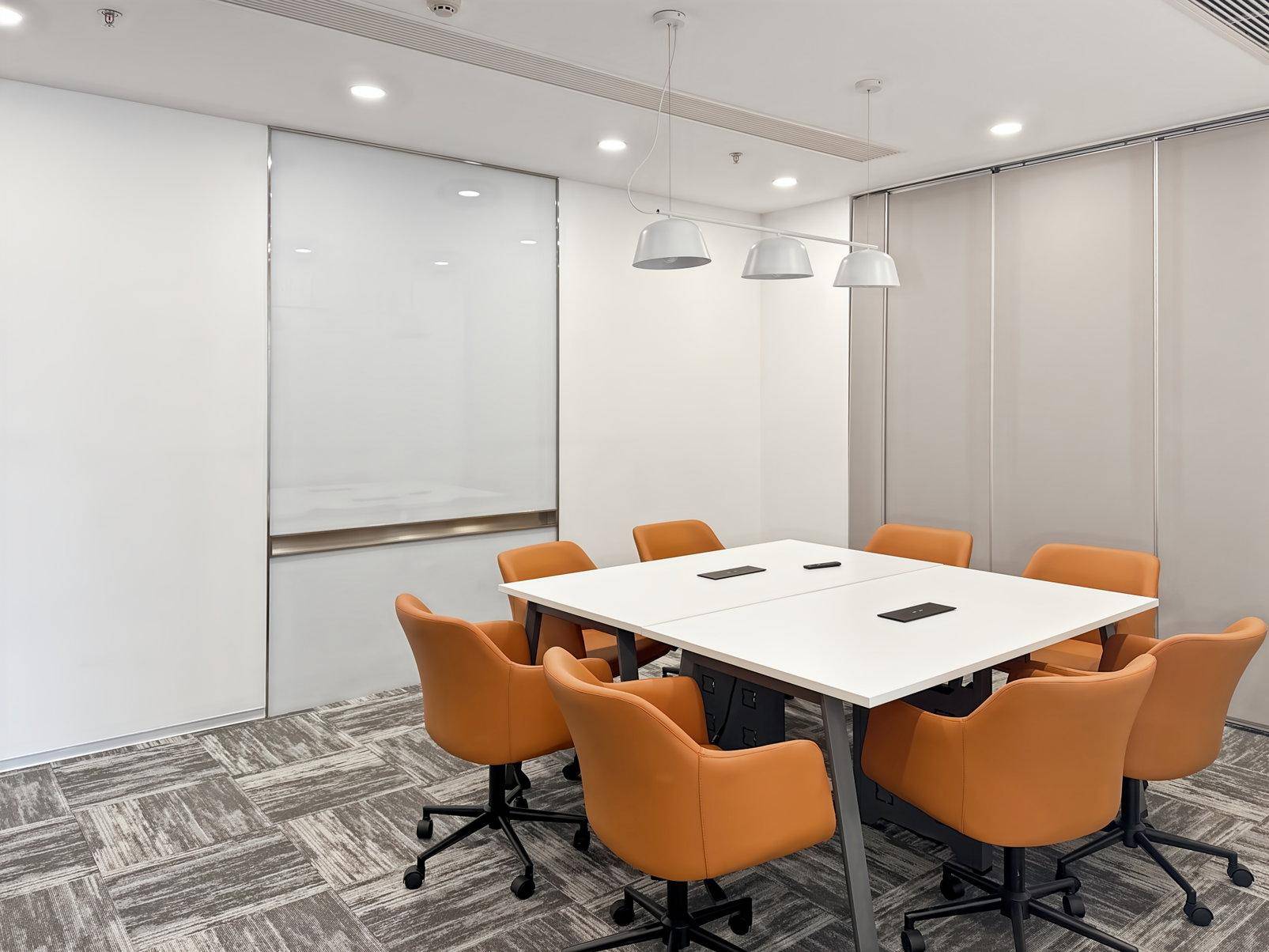 Heller Konferenzraum mit großem weißen Tisch, bequemen orangefarbenen Stühlen und einer Whiteboard-Wand.
