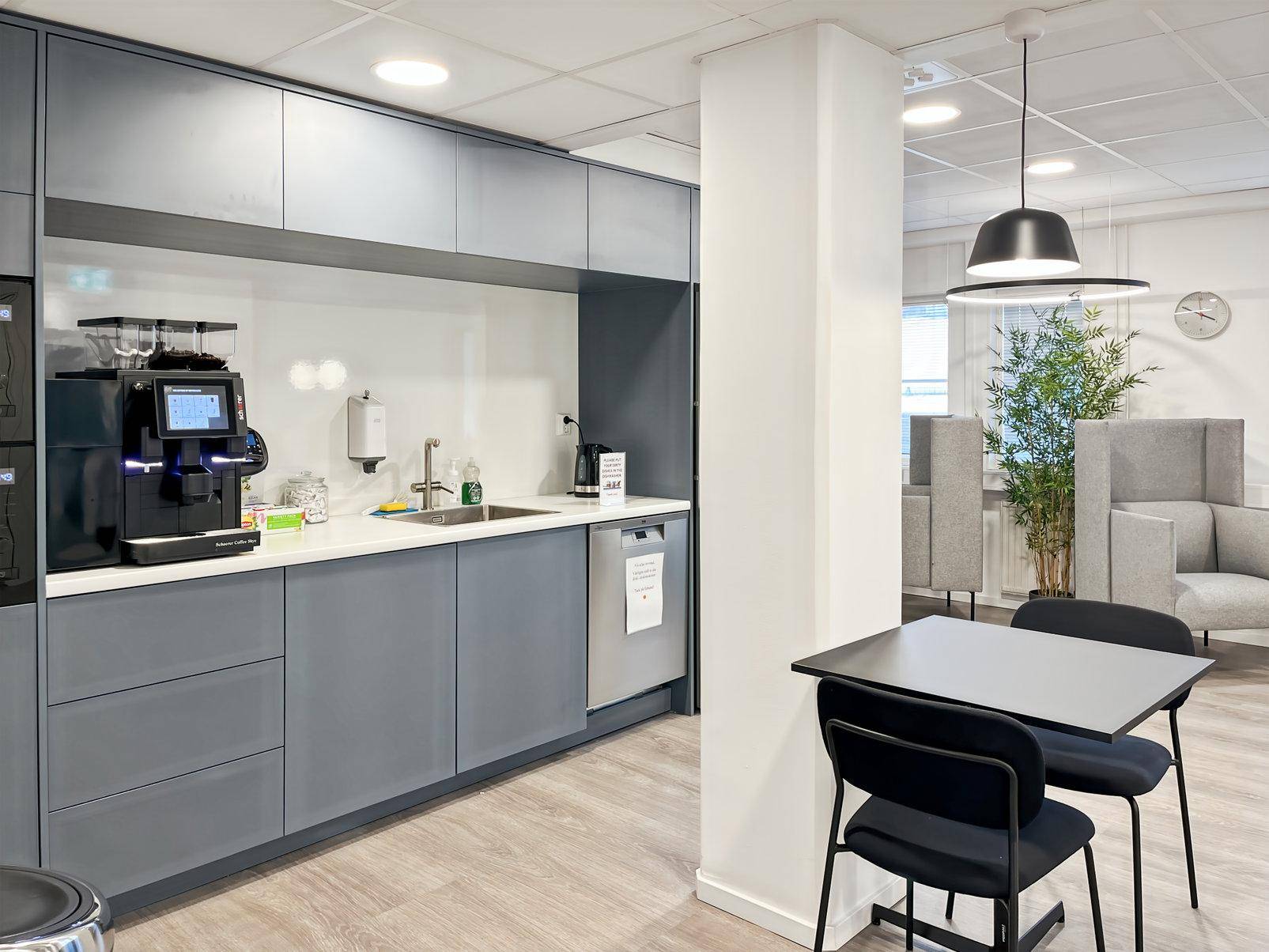 Moderne Büroküche mit grauen Schränken, weißer Arbeitsplatte, Spülmaschine und Kaffeemaschine, inklusive Essbereich.
