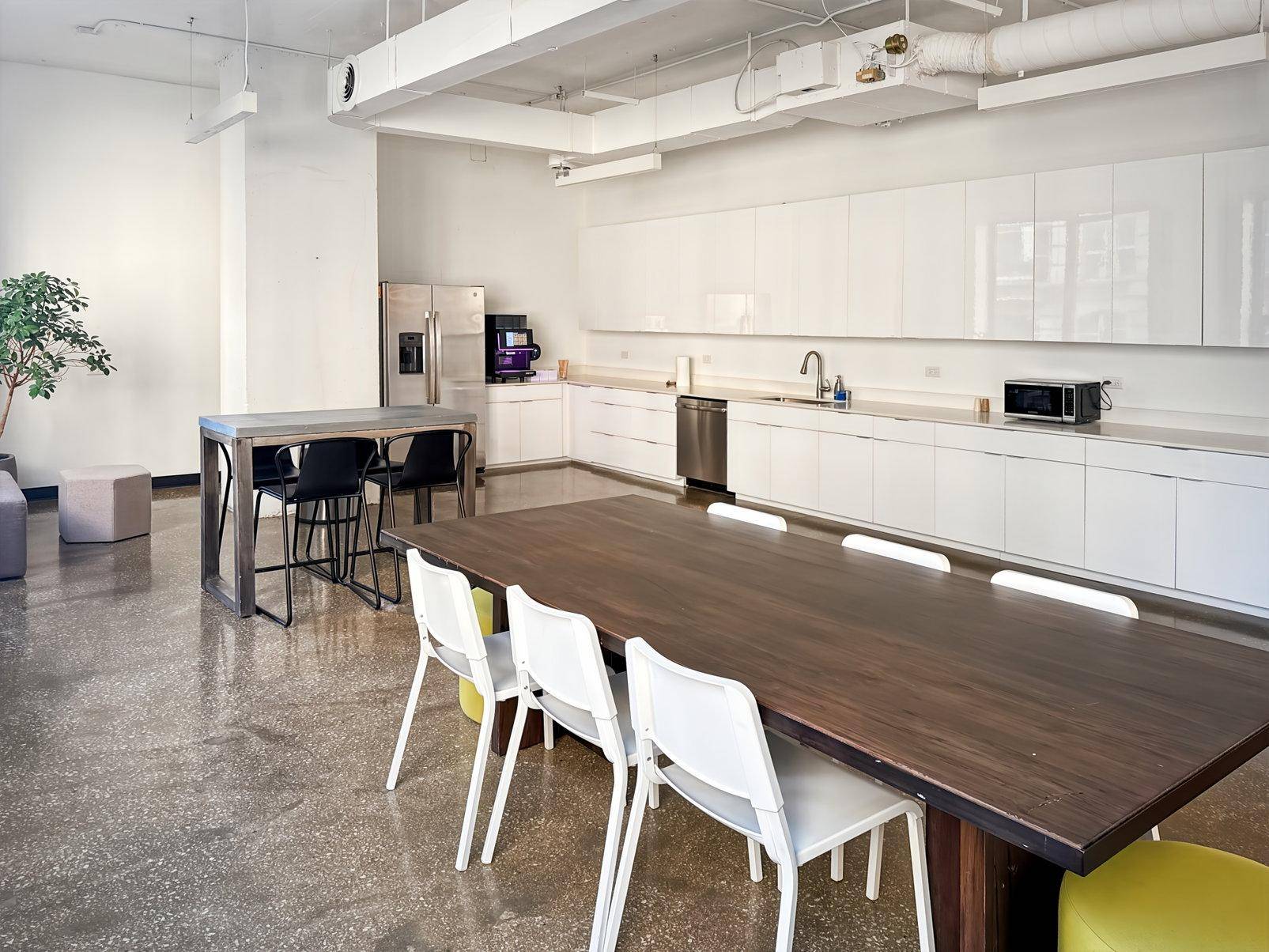 Große moderne Büroküche mit weißen Hochglanzschränken, Edelstahlofen und Essbereich mit langem Holztisch.
