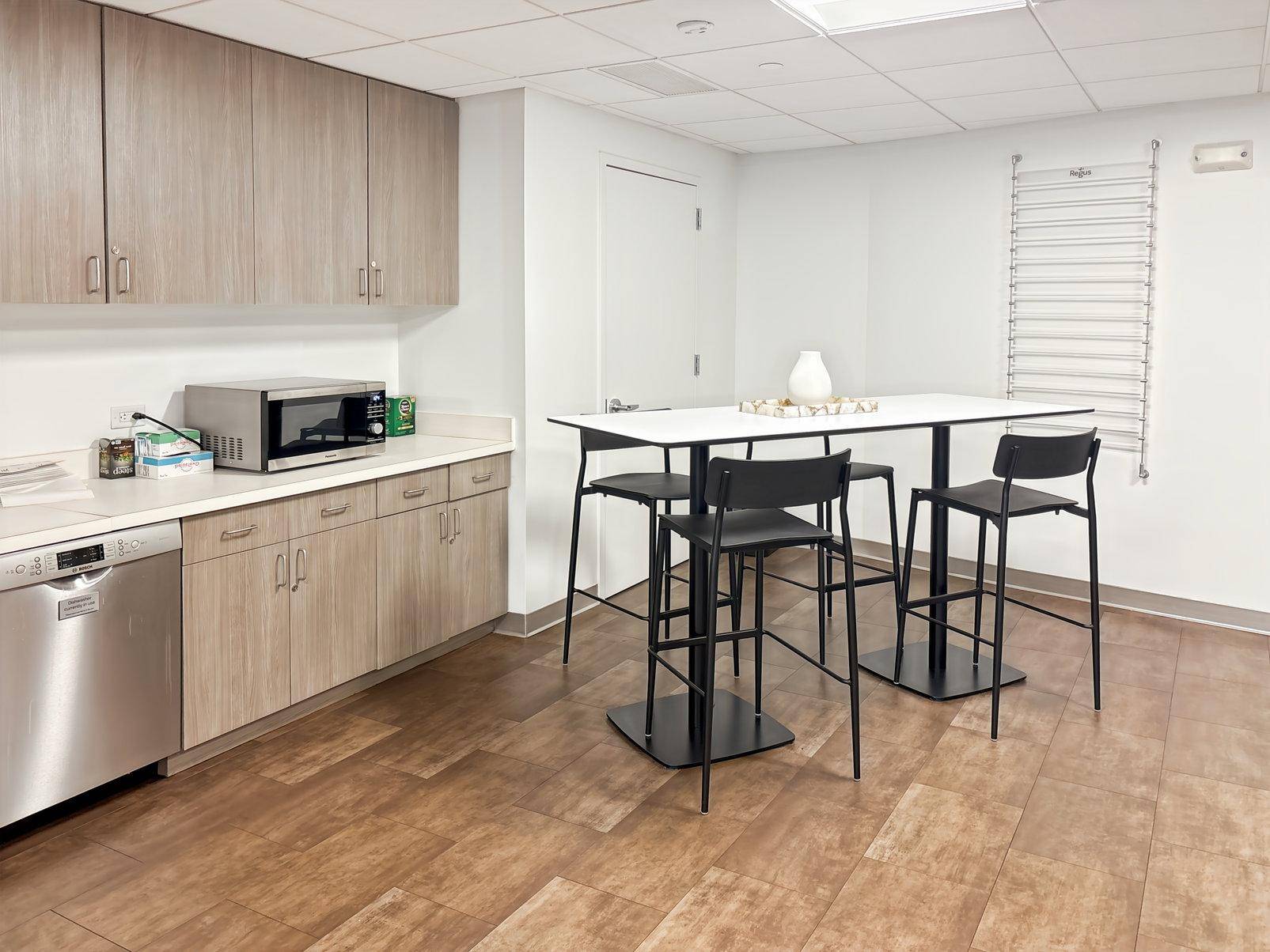 Funktionale Büroküche mit Holzschränken, Mikrowelle, Geschirrspüler und Stehtisch mit Barhockern.