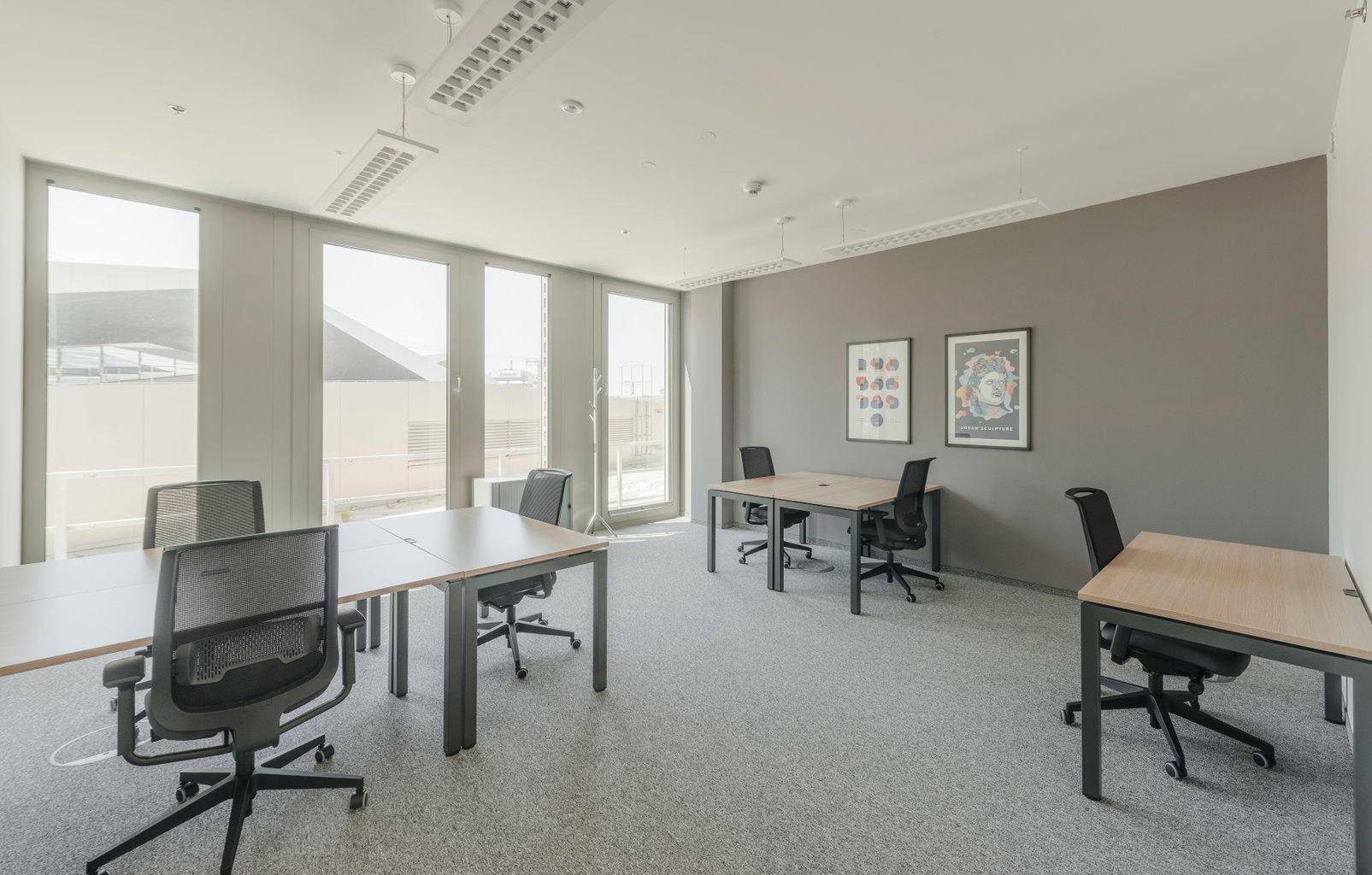 Helles Büro mit mehreren Arbeitsplätzen, grauen Wänden und großen Fenstern für viel Tageslicht.