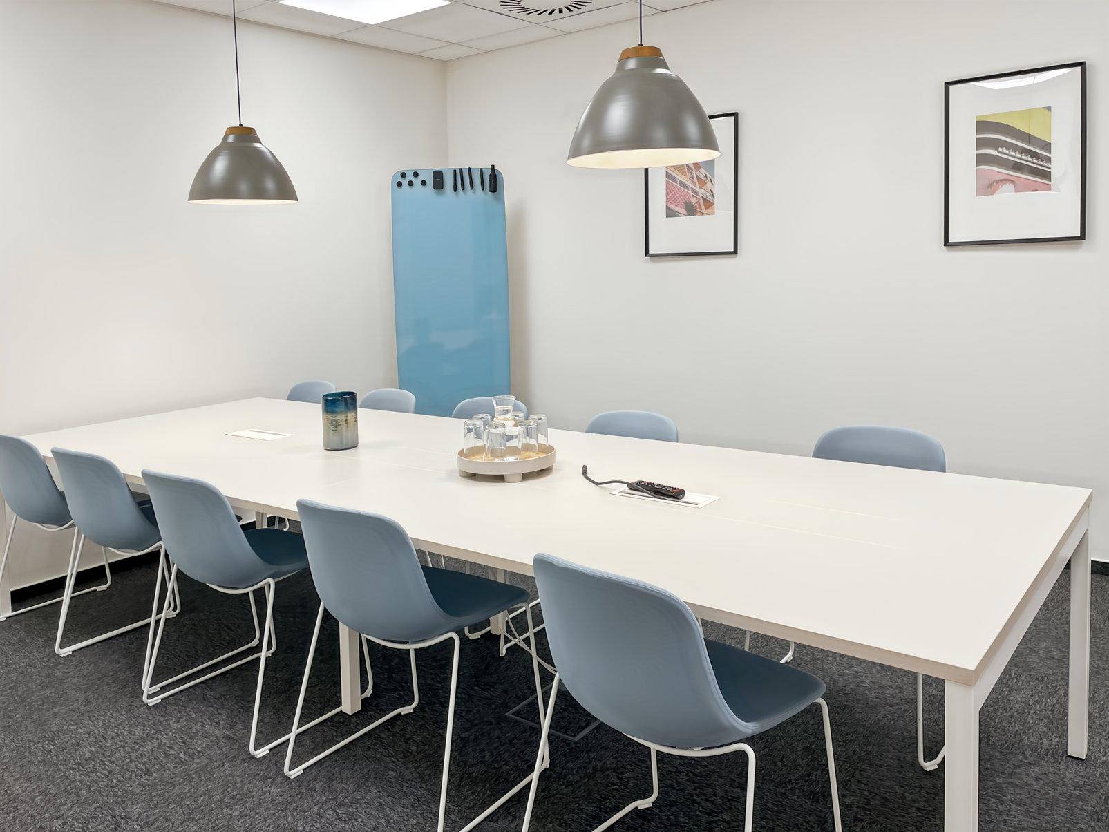 Moderner Konferenzraum mit einem langen weißen Tisch, hellblauen Stühlen und zwei stilvollen Pendelleuchten.