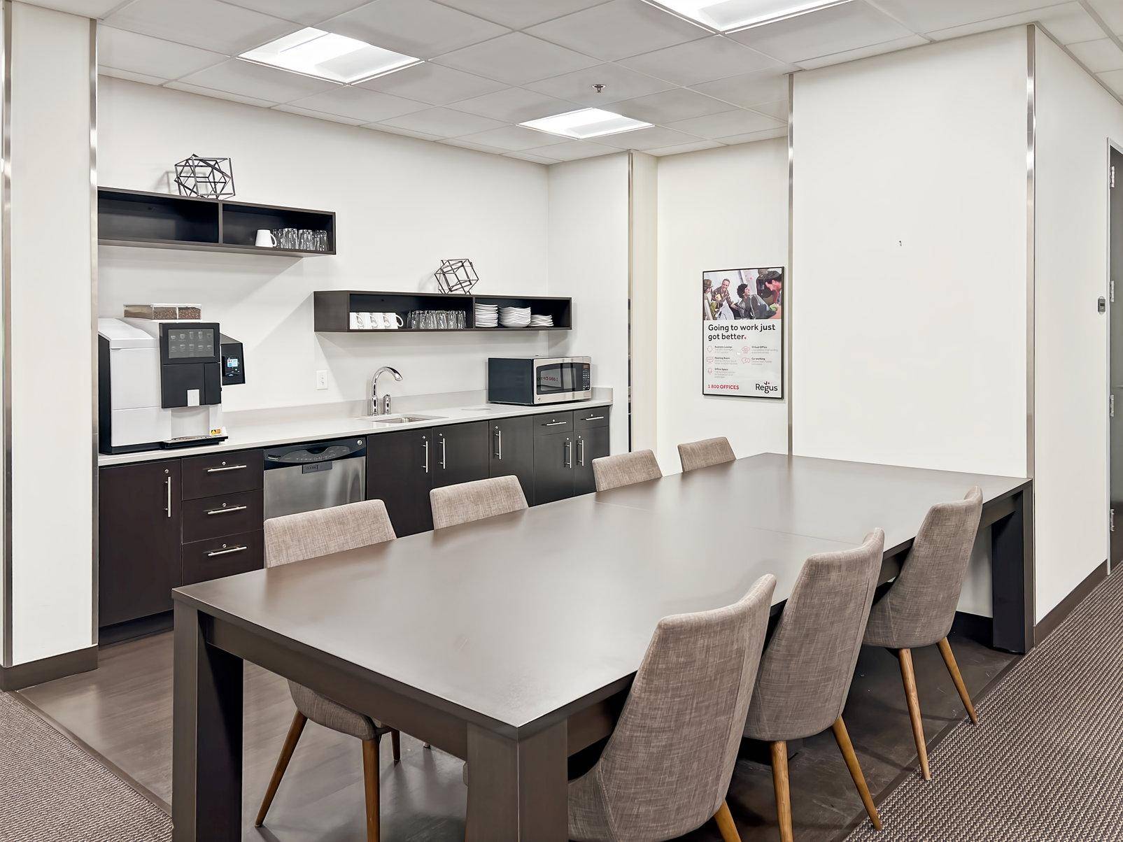 Moderne Büroküche mit langer Tafel, dunklen Schränken, Spülmaschine und Kaffeemaschine für Mitarbeiter.
