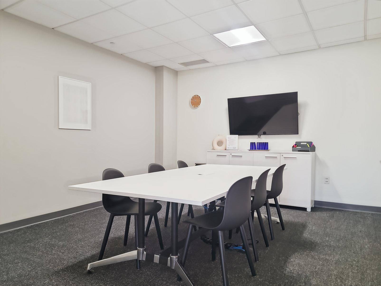 Kleiner Besprechungsraum mit weißem Tisch, schwarzen Stühlen und einem großen Wandfernseher für Präsentationen.