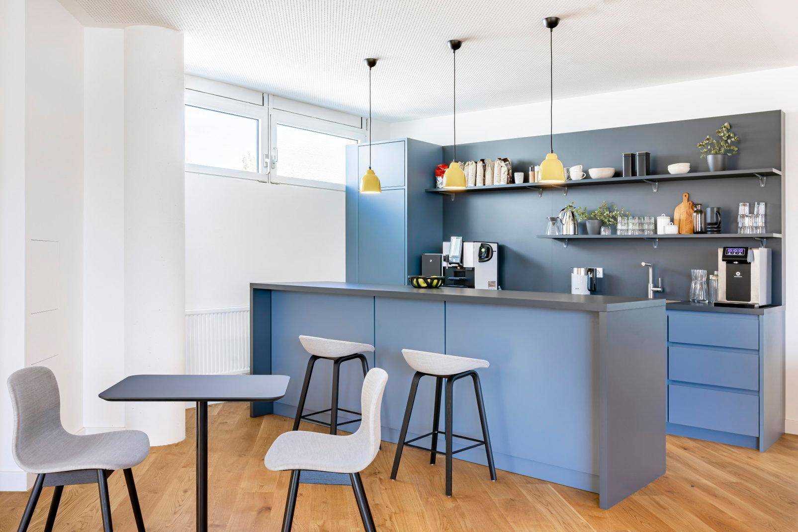 Moderne Teeküche mit blauen Schränken, Holzboden und einem kleinen Essbereich mit Fenstern.