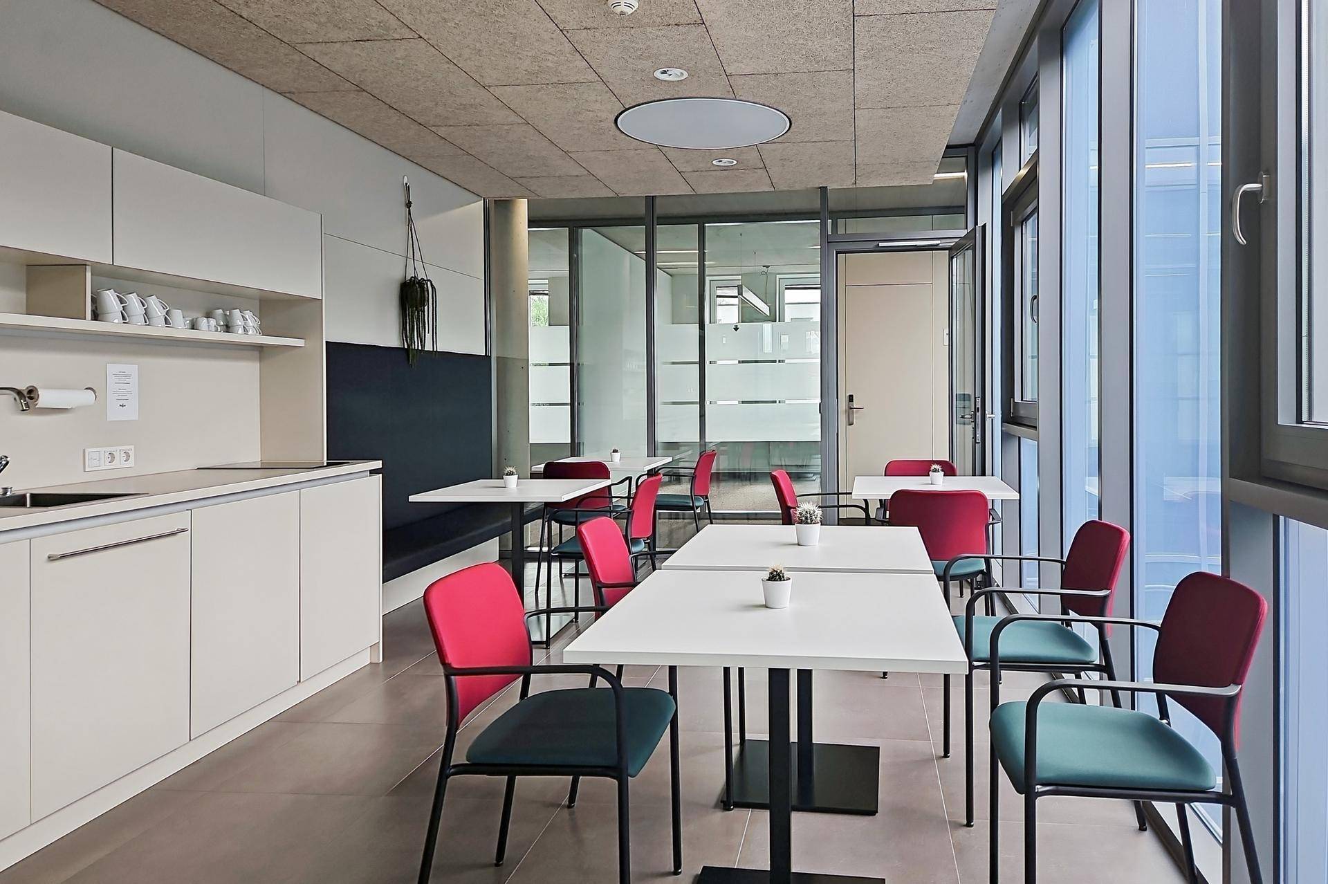 Moderner Pausenraum mit kleiner Küchenzeile, mehreren Tischen und Stühlen sowie großen Fenstern.
