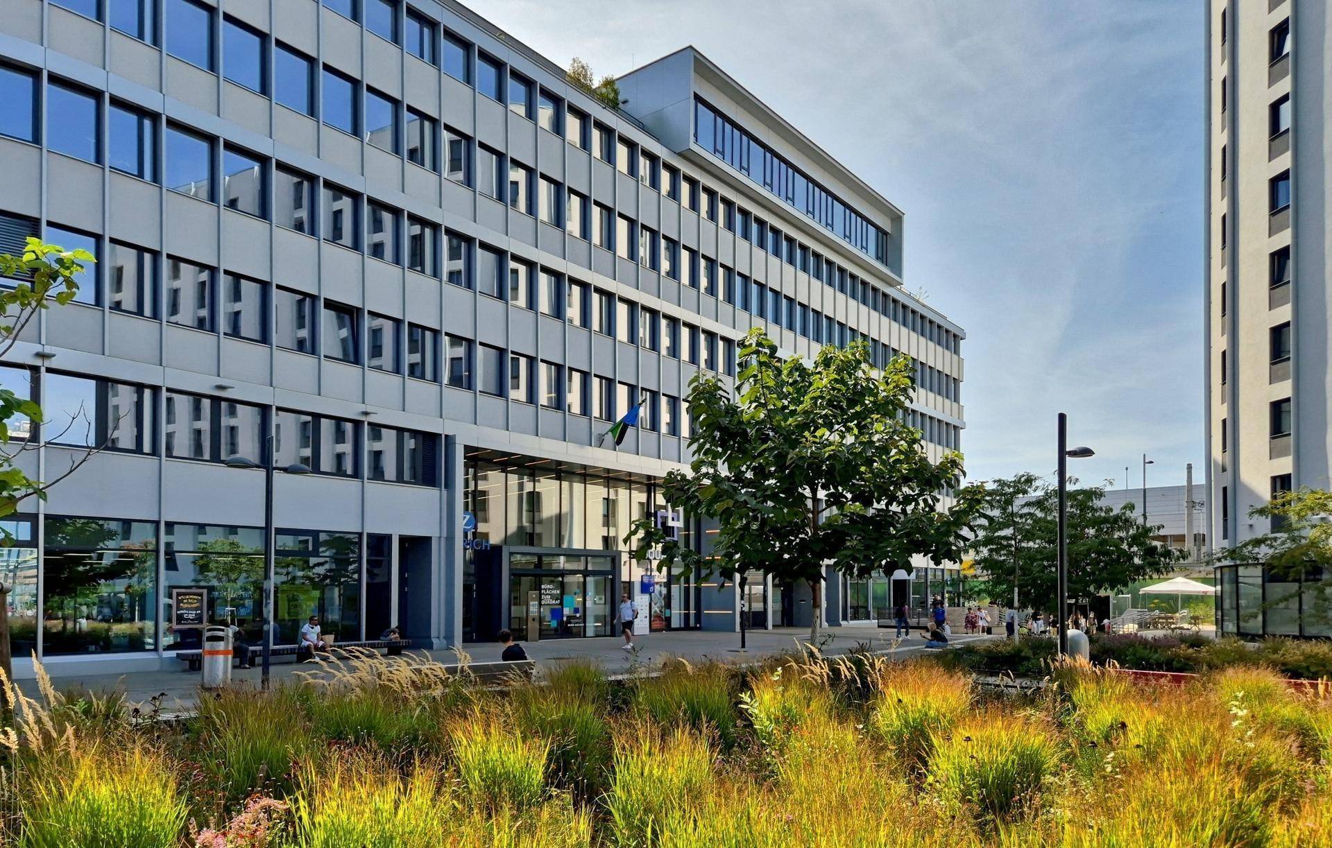Modernes Bürogebäude mit Glasfassade und umliegender Grünfläche unter blauem Himmel.