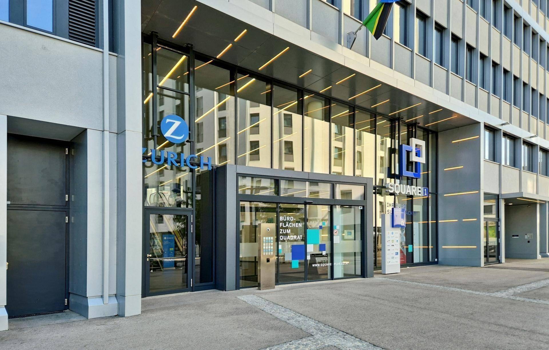 Moderner Haupteingang eines Bürogebäudes mit großer Glasfront und automatischen Türen, gut beleuchtet.