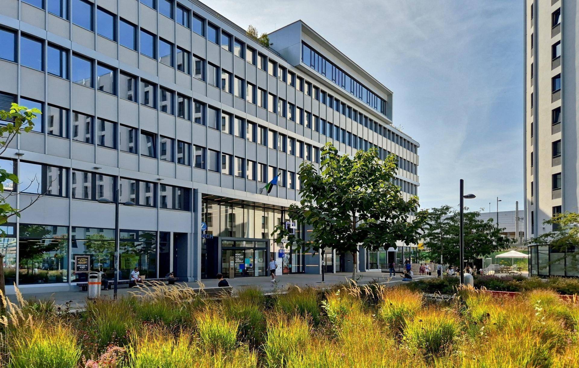 Modernes Bürogebäude mit Glasfassade und gepflegter Grünanlage im Vordergrund unter blauem Himmel.