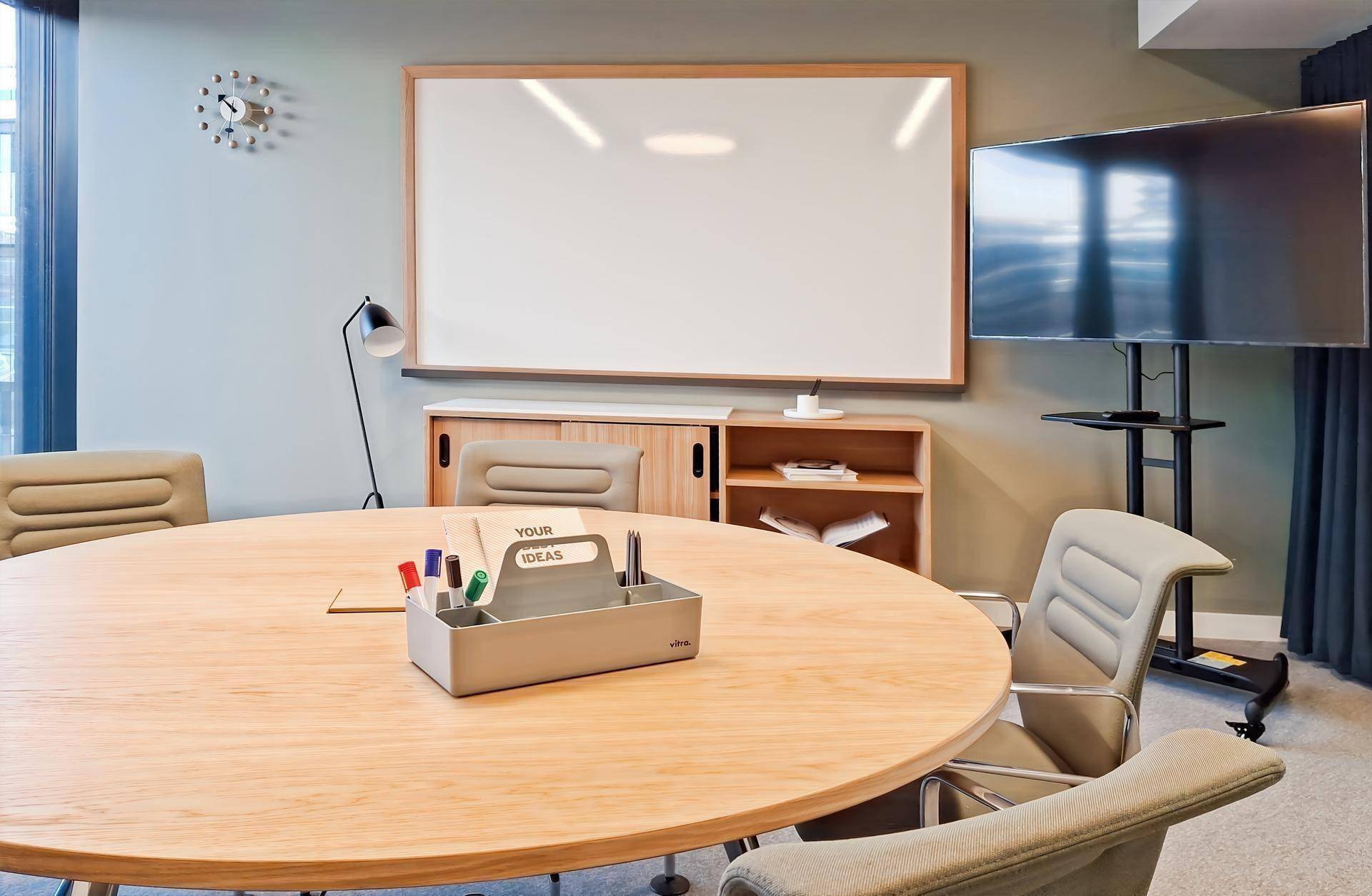 Nahaufnahme eines runden Konferenztisches mit Utensilien, Whiteboard und Bildschirm im Hintergrund.
