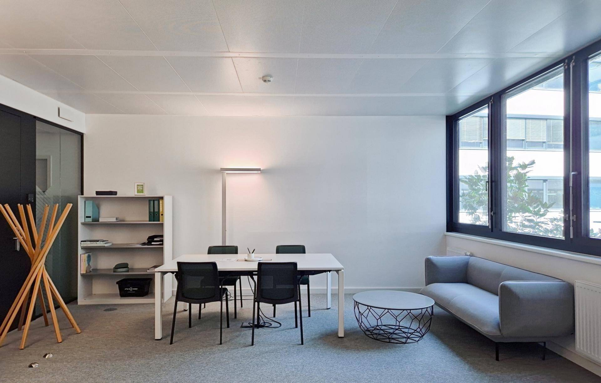 Funktionaler Büroraum mit Besprechungstisch, Sofa, Regalen und großen Fenstern für viel Tageslicht.