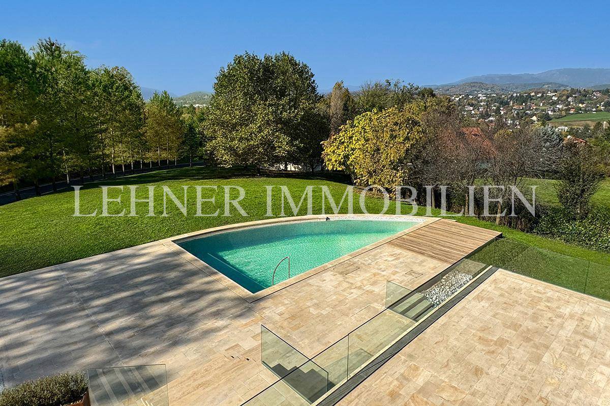 Geschwungener Swimmingpool mit Holzdeck und weitläufiger Steinterrasse, umgeben von grüner Landschaft.