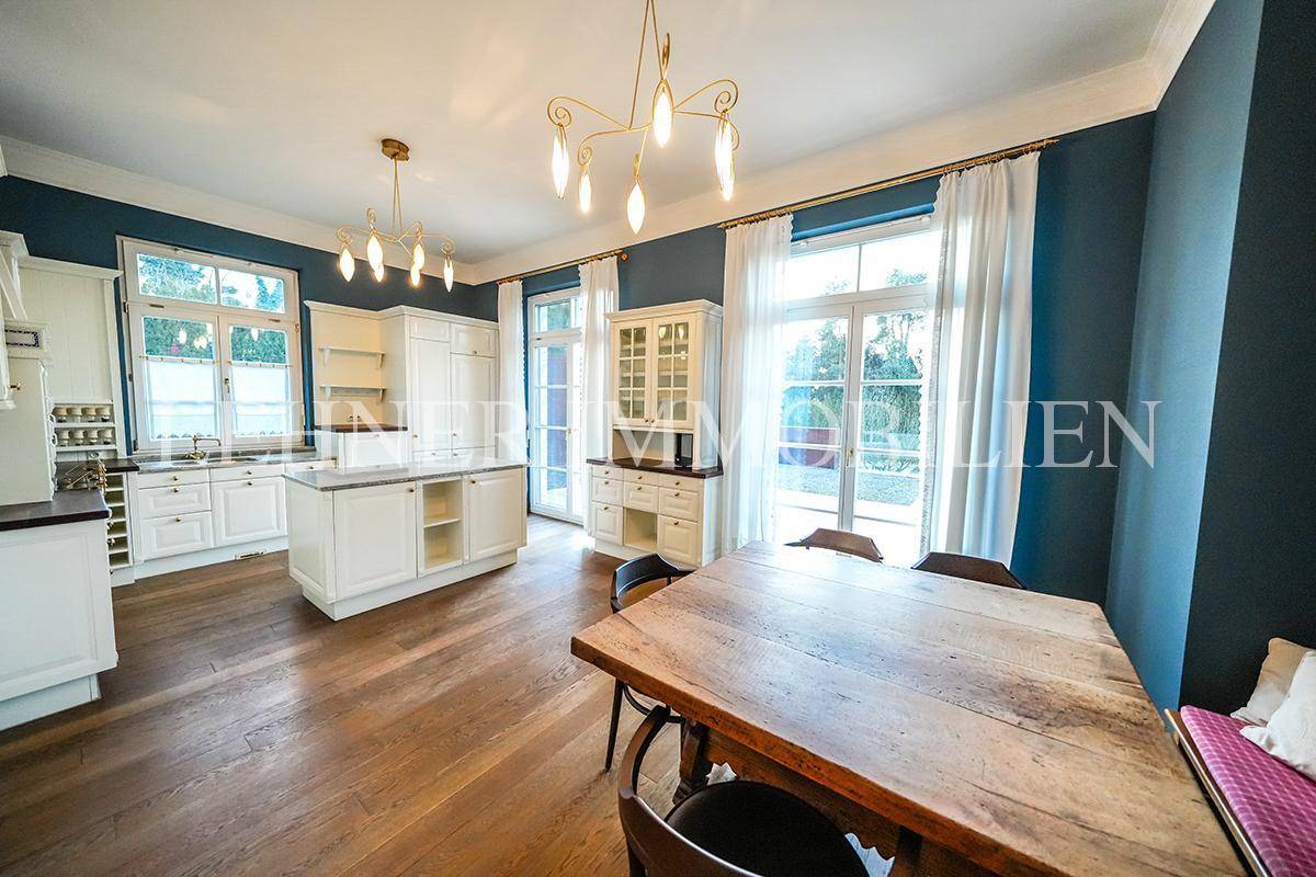 Großzügige Wohnküche mit weißen Schränken, Kücheninsel und Essbereich mit Blick ins Grüne.