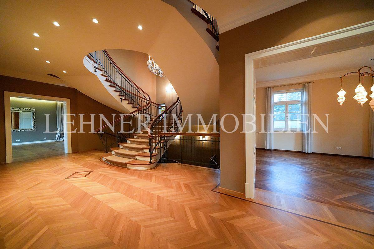 Eindrucksvoller Flur mit Fischgrätparkett, hohen Decken und Blick auf das geschwungene Treppenhaus.