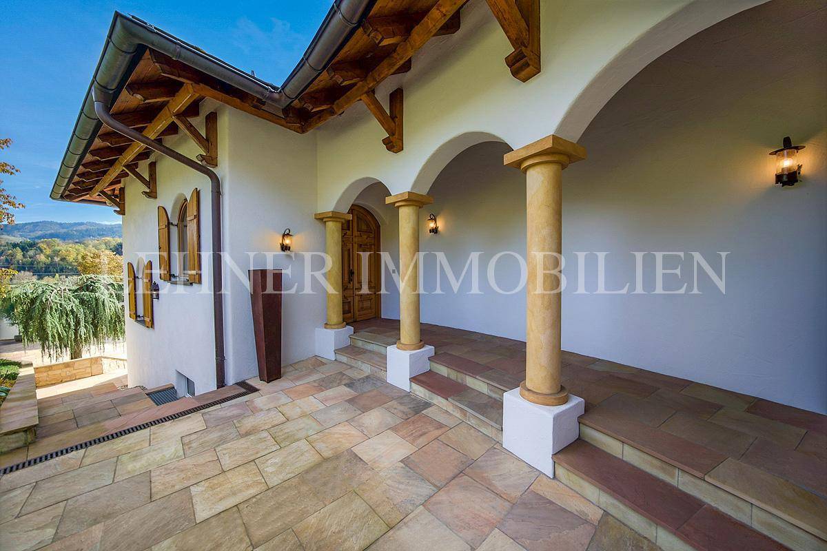 Überdachter Eingangsbereich mit Säulen und Holztür, gepflastertem Boden und Außenleuchten.