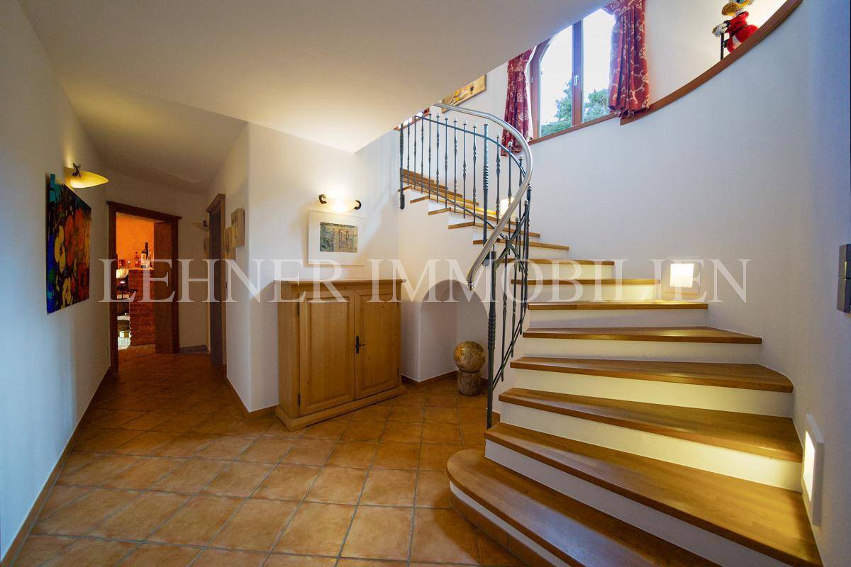 Eingangsbereich mit geschwungener Holztreppe, schmiedeeisernem Geländer und Terrakottafliesen.