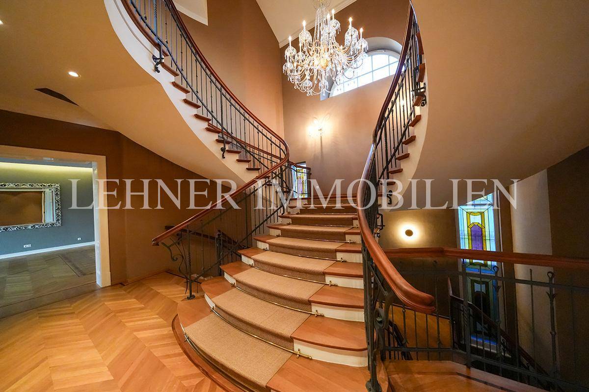Elegantes Treppenhaus mit geschwungener Treppe, Kronleuchter und farbigem Glasfenster.