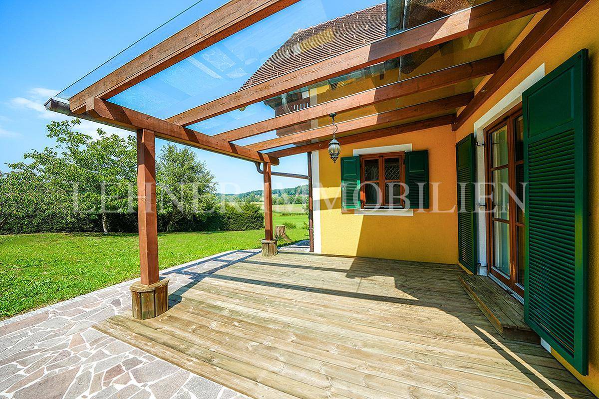 Sonnige Holzterrasse mit Glasüberdachung und Zugang zum Garten, umgeben von Natur.