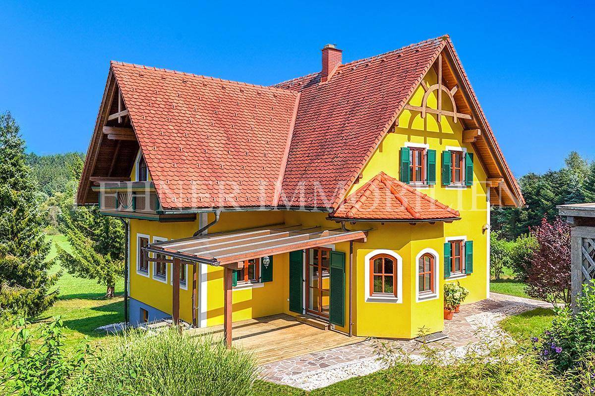 Idyllisches Einfamilienhaus mit gelber Fassade, grünen Fensterläden und überdachter Terrasse in sonniger Lage.