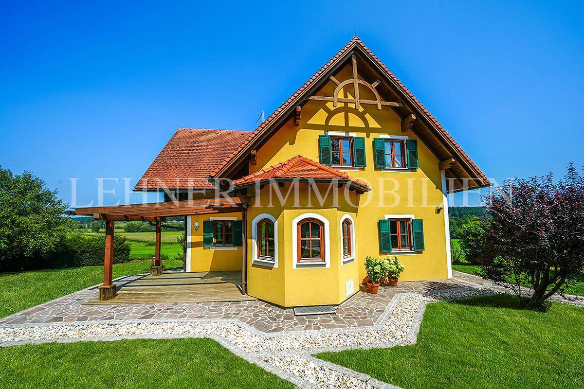 Attraktives Landhaus mit gelber Fassade, grünen Fensterläden und überdachter Holzterrasse, umgeben von Natur.