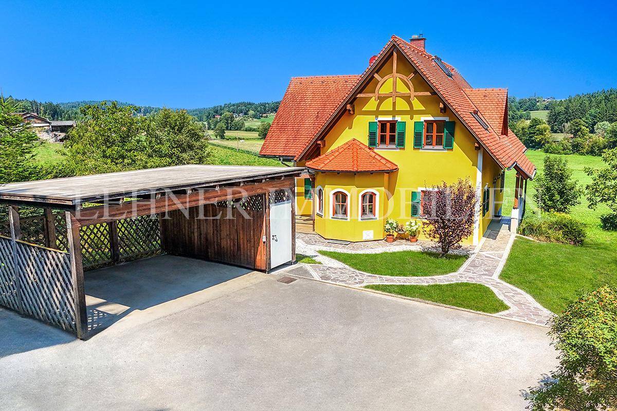 Landhaus mit gelber Fassade, rotem Dach und separatem Holzcarport, umgeben von grüner Landschaft.